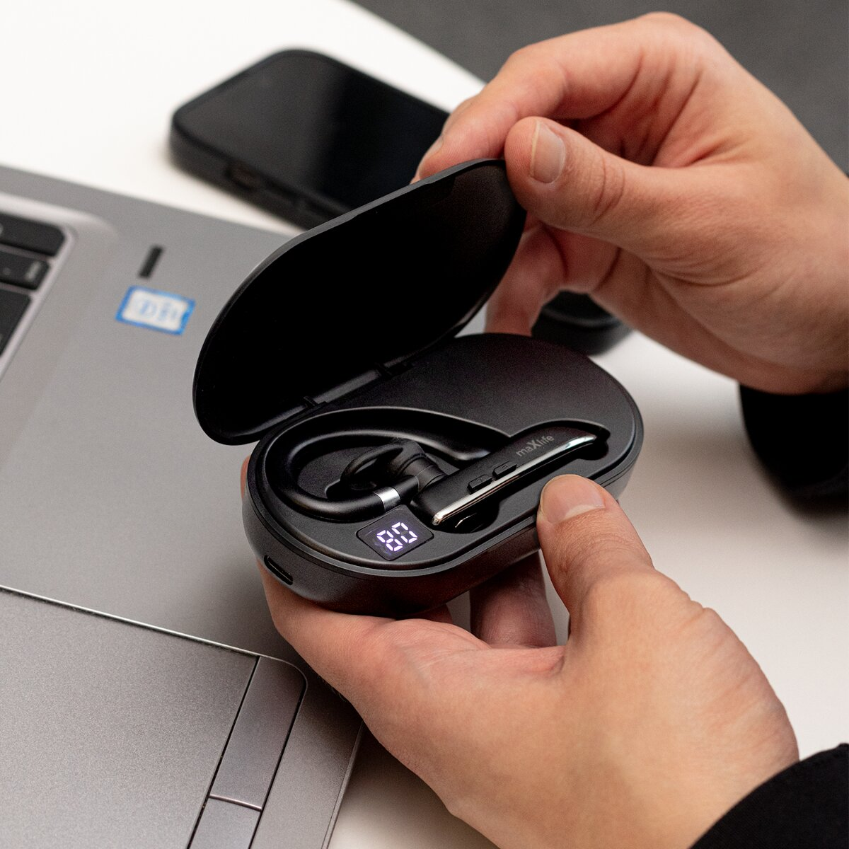 Mani che tengono un auricolare nero nel suo case di ricarica. Laptop e telefono dietro.