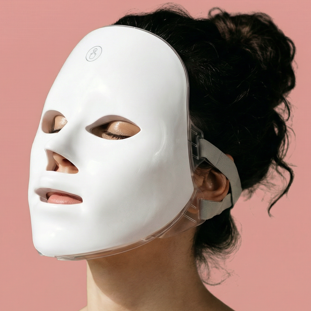 Biała maska LED na twarz osoby z ciemnymi włosami, na różowym tle.