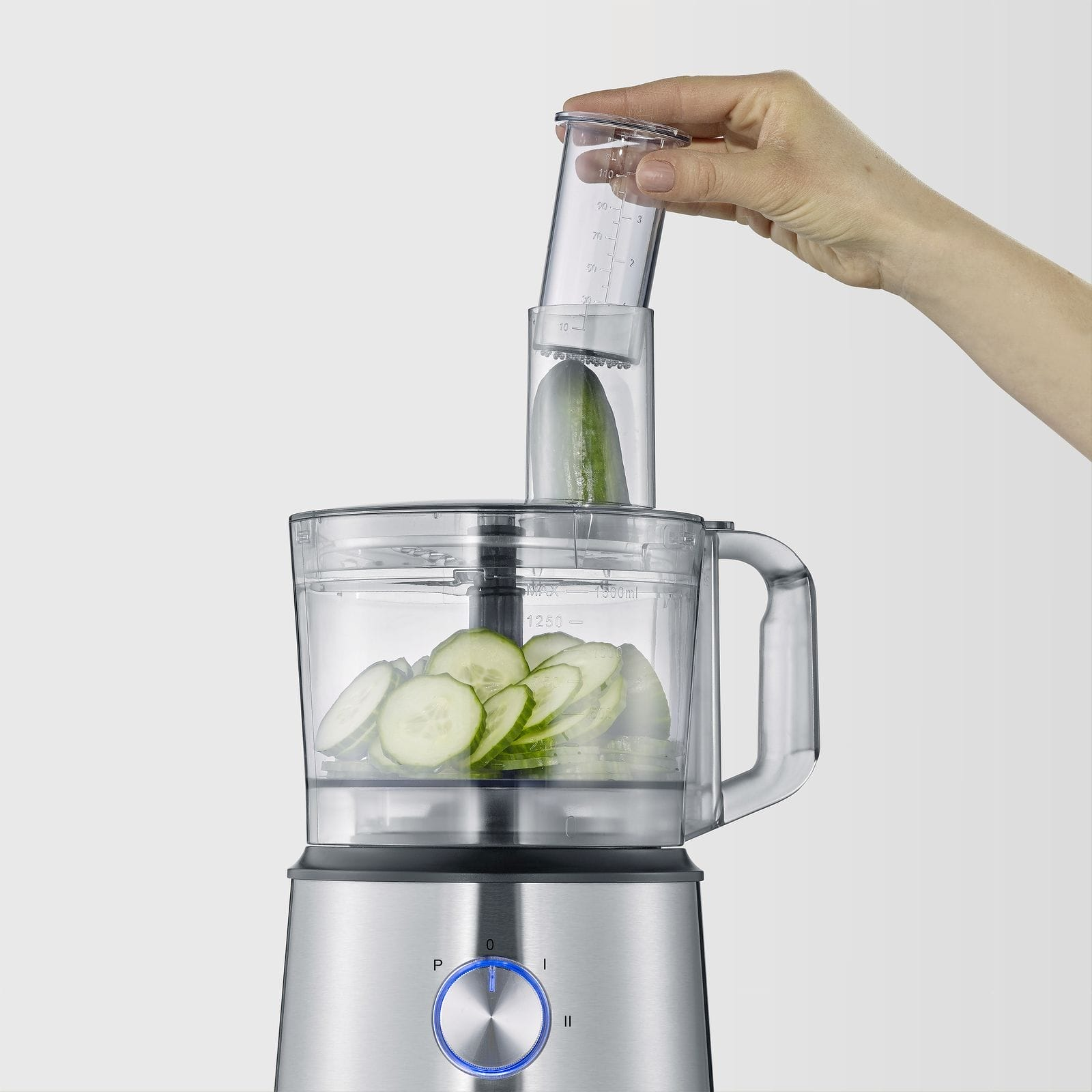 Een hand plaatst een komkommer in een keukenmachine-opzetstuk met schijfjes.