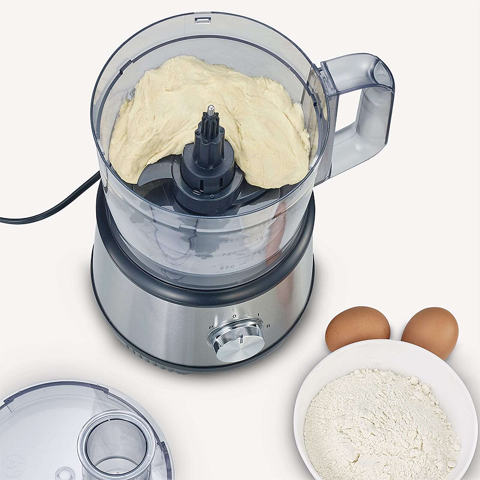 Keukenmachine met deeg erin, met bloem en eieren ernaast.