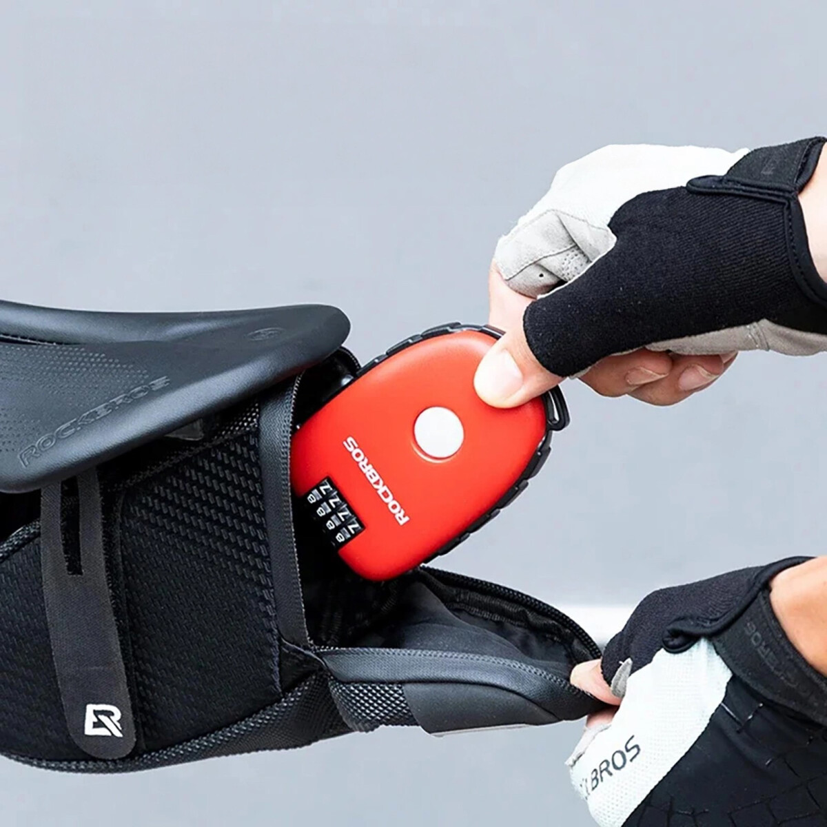 Eine Hand mit Handschuh legt ein rotes Zahlenschloss in eine Fahrradtasche unter einem Sattel.