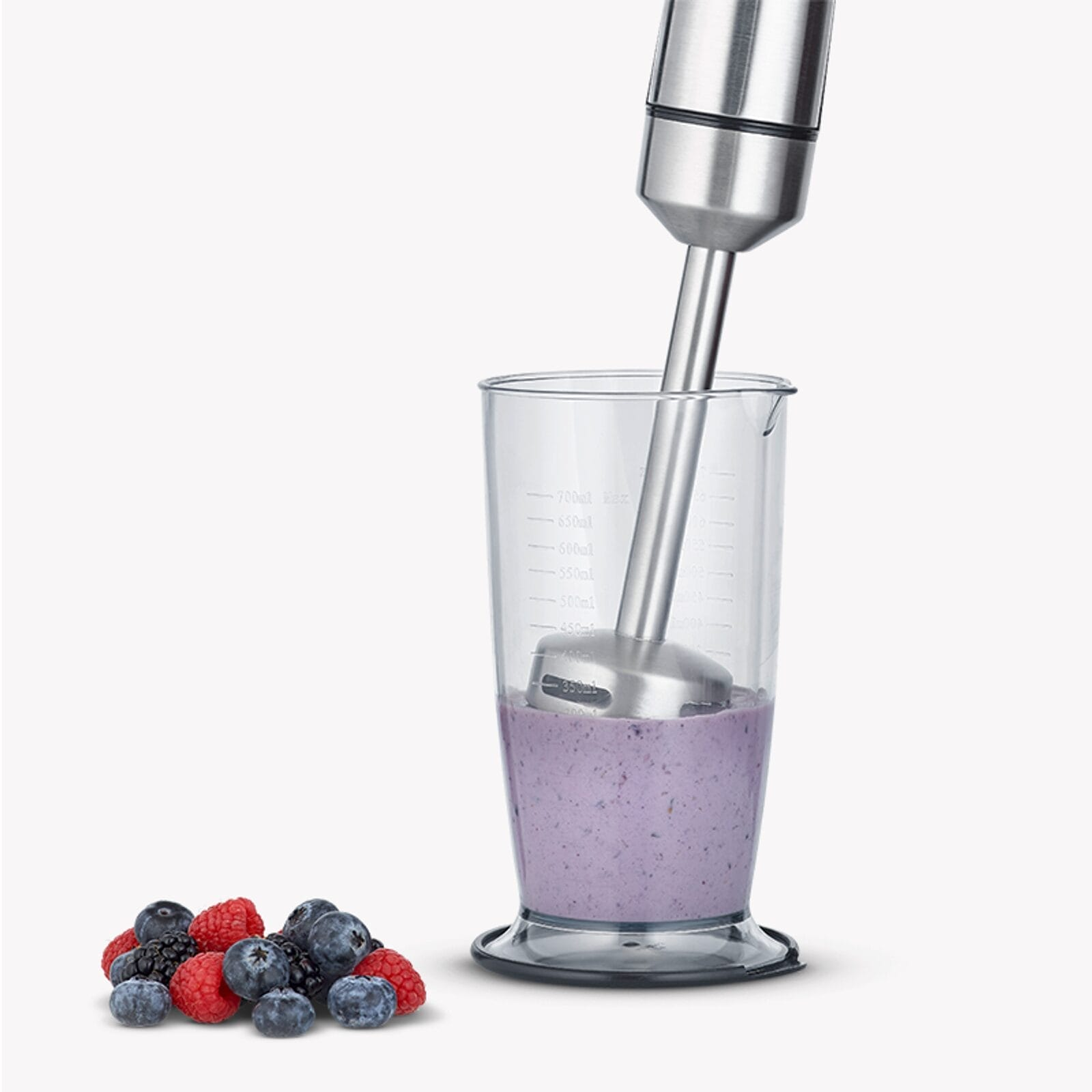 A hand blender mixes berries in a measuring cup. Berries are next to it.