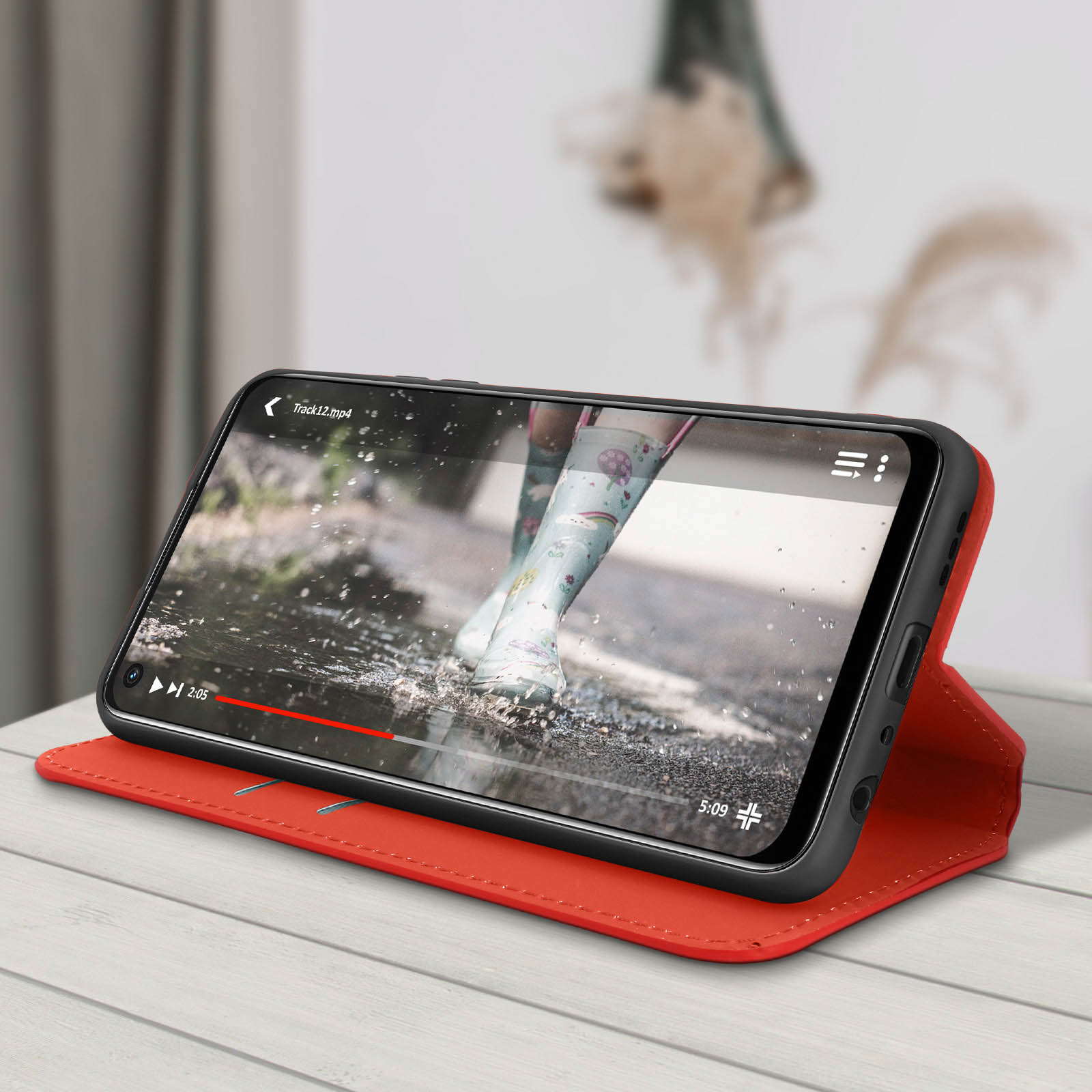 Een smartphone in een rode hoes staat op een houten tafel en toont een video van voeten in regenlaarzen die in water spatten.
