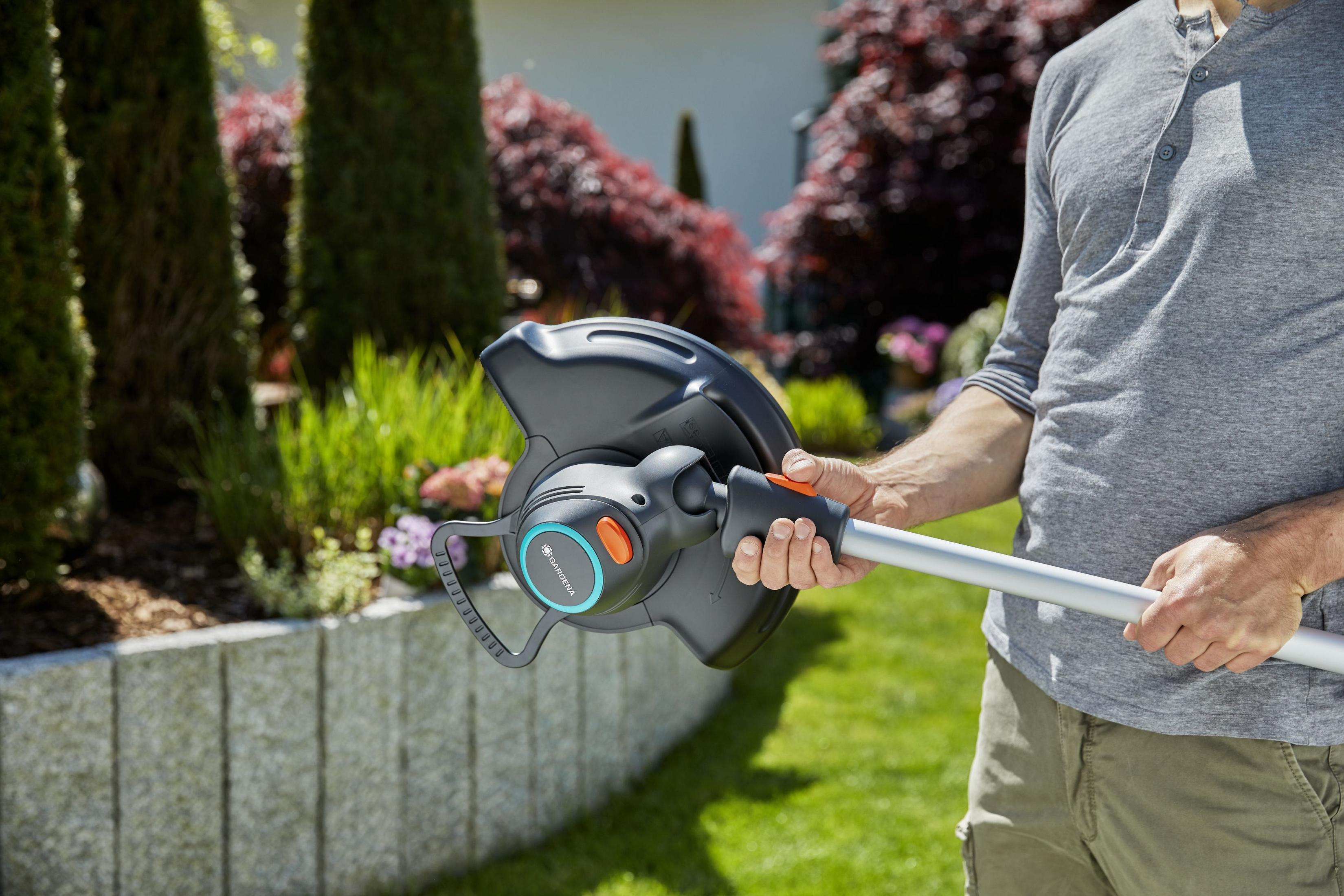 Mann hält einen Rasentrimmer im Garten, mit Bäumen und Blumen im Hintergrund.