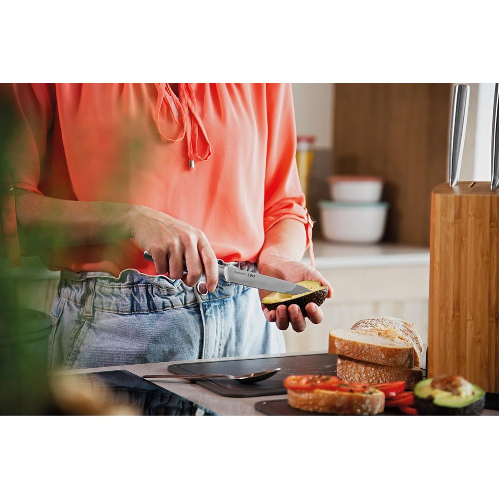 Persoon snijdt avocado met mes in keuken. Brood en tomaten ernaast.
