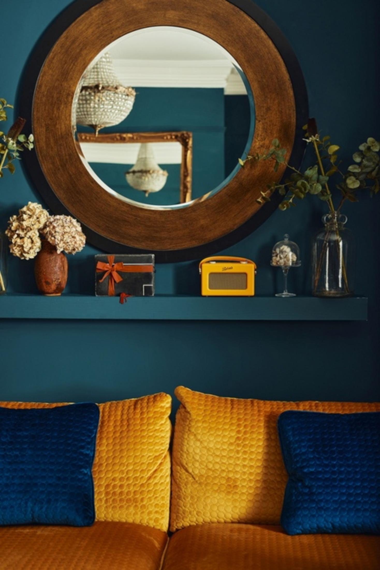 Okrągłe lustro nad półką z dekoracjami. Poniżej sofa z poduszkami.