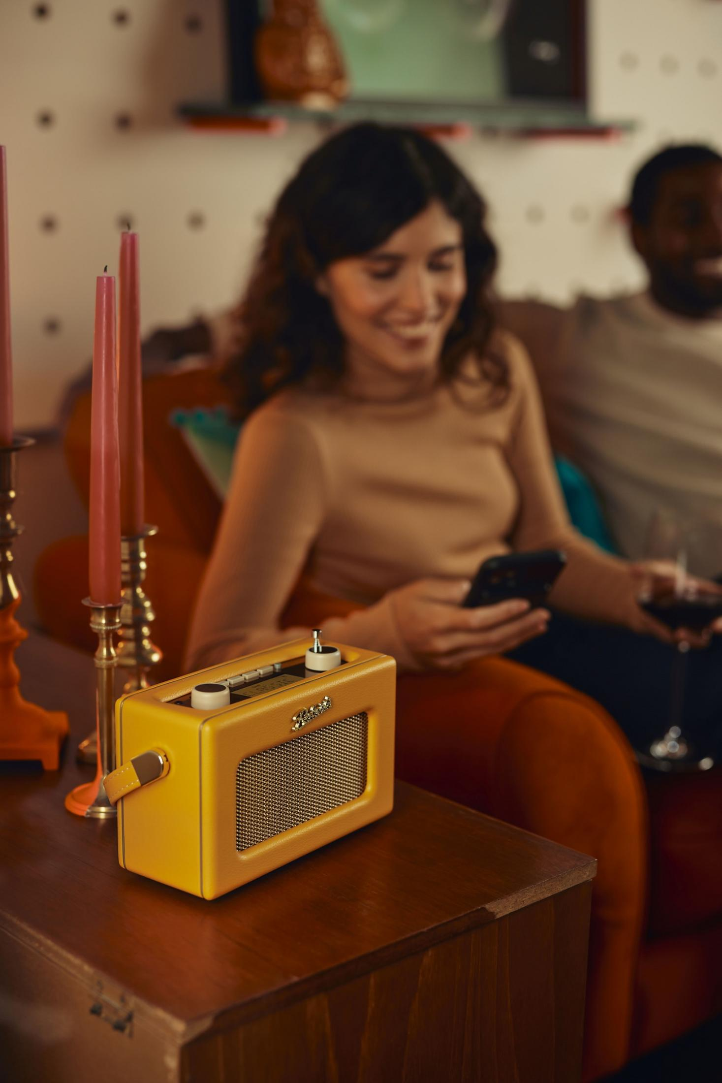 Ein gelber Retro-Radio steht auf einem Holztisch, ein Paar sitzt im Hintergrund.