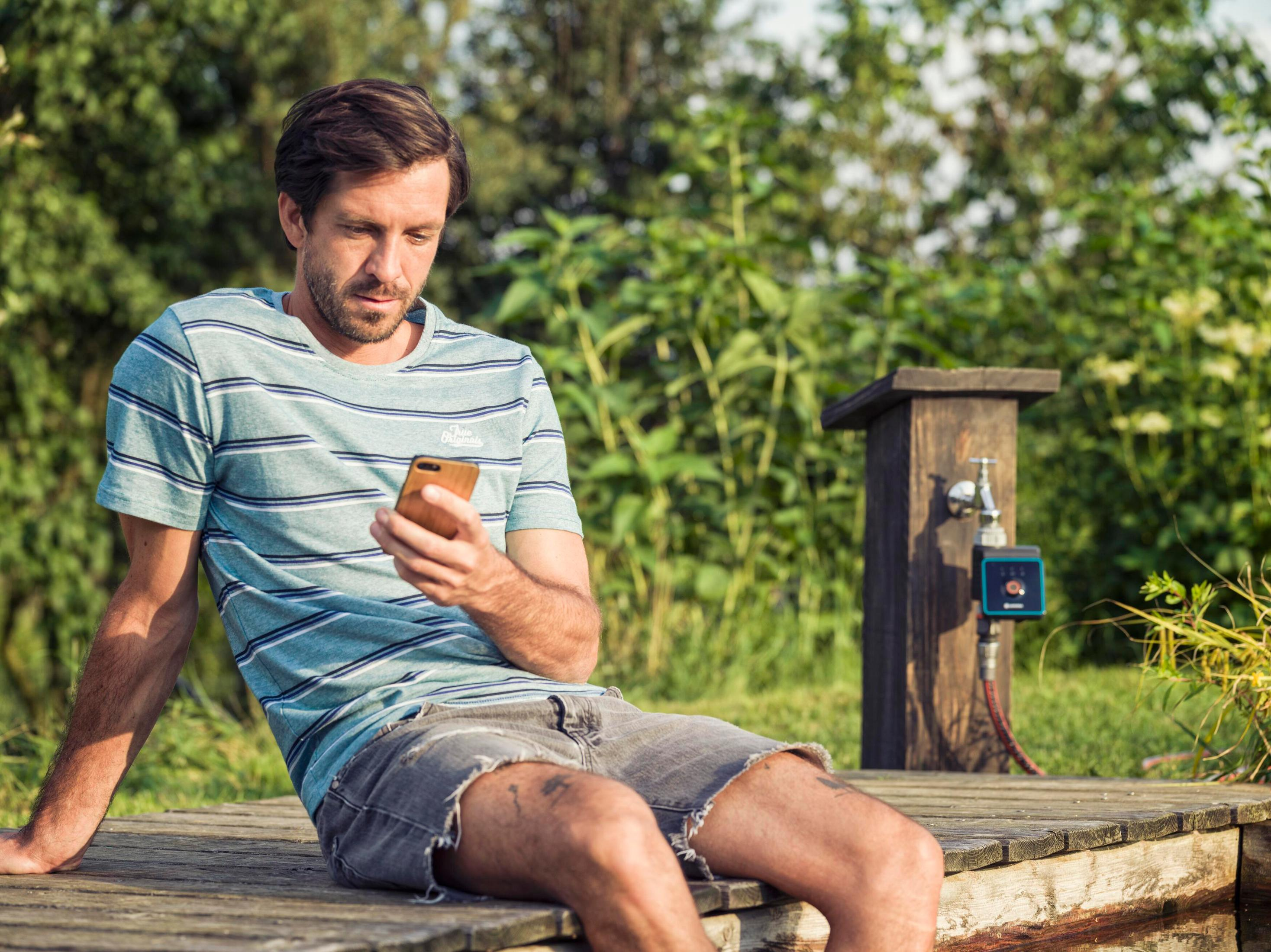 Ein Mann sitzt auf einer Holzplattform und schaut auf sein Handy. Im Hintergrund sind Gräser und ein Wasserhahn.
