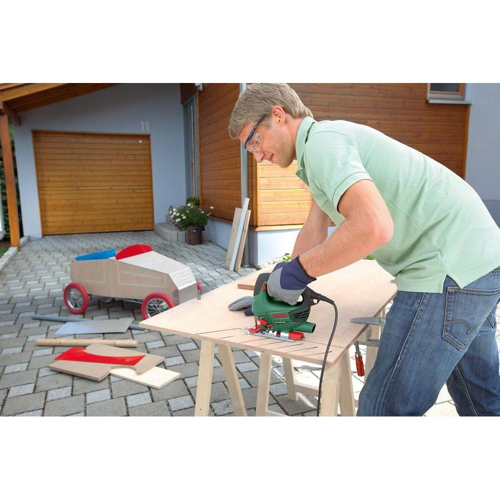 Mężczyzna w okularach używa wyrzynarki do drewna; samochodzik z tyłu.