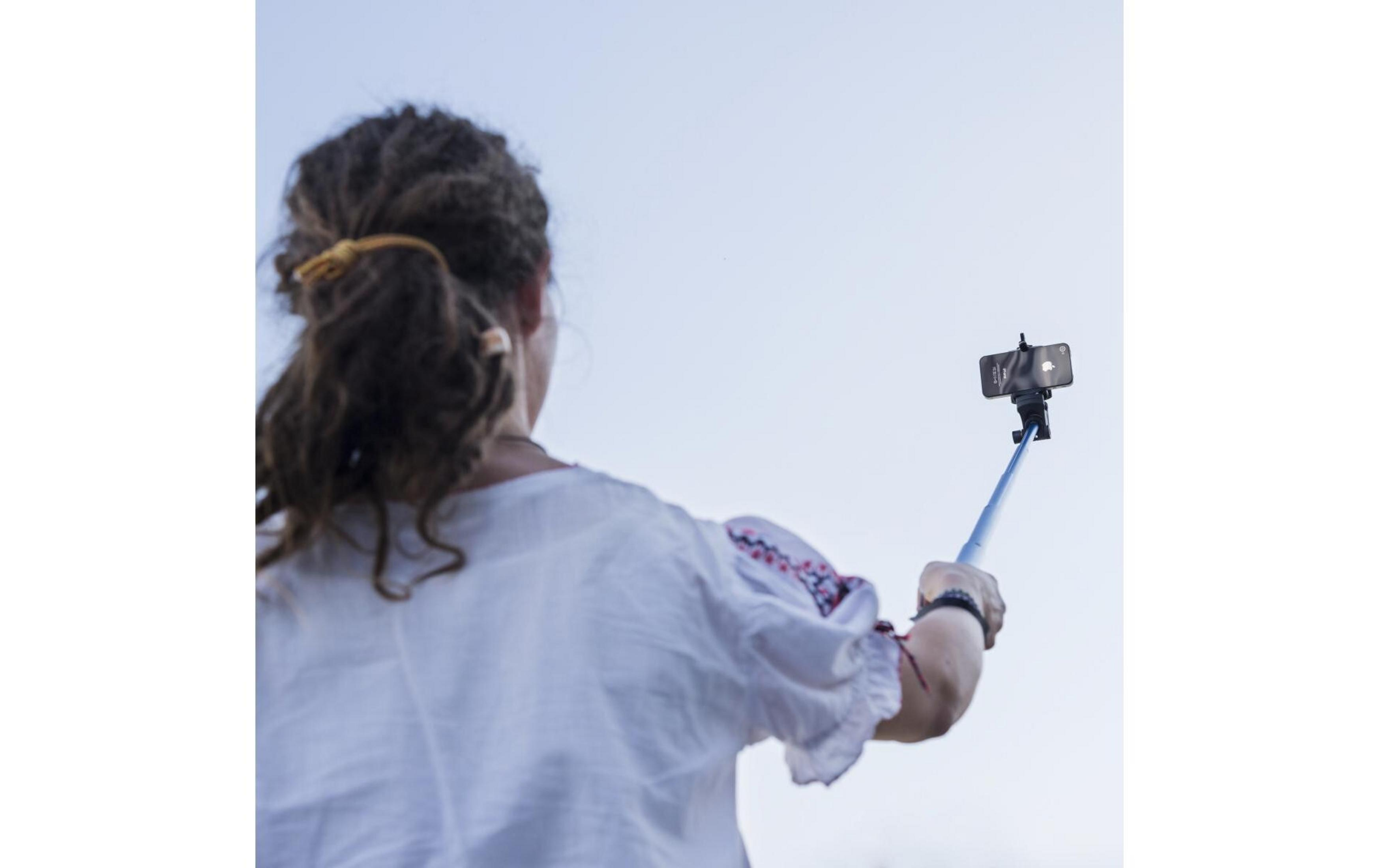 Frau mit Dreadlocks in weißem Oberteil macht Selfie mit Stick gegen blauen Himmel.