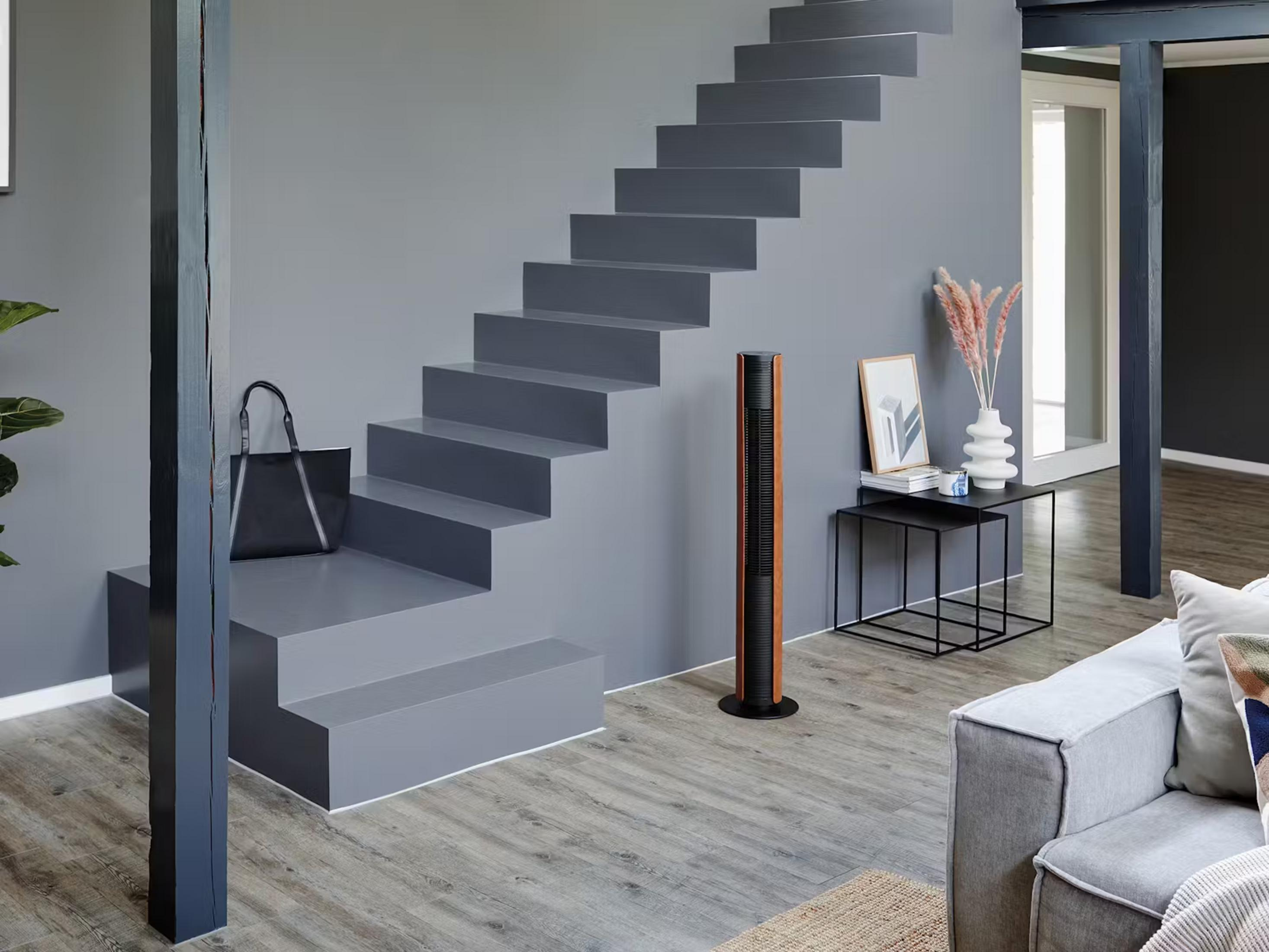 Un espace de vie moderne avec escalier gris, ventilateur tour, tables d'appoint et canapé.