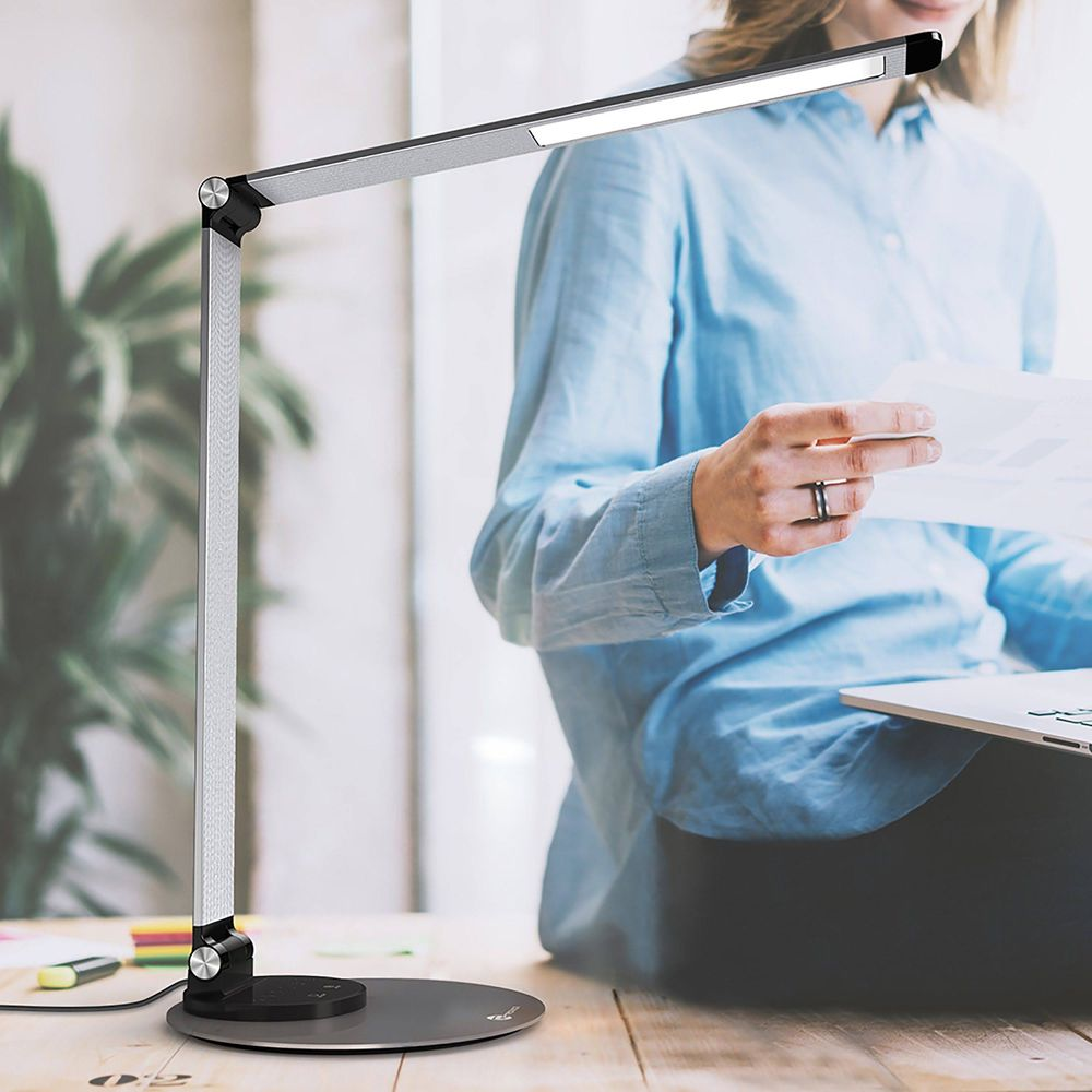 Eine Frau im blauen Hemd liest Dokumente neben einer modernen Schreibtischlampe.