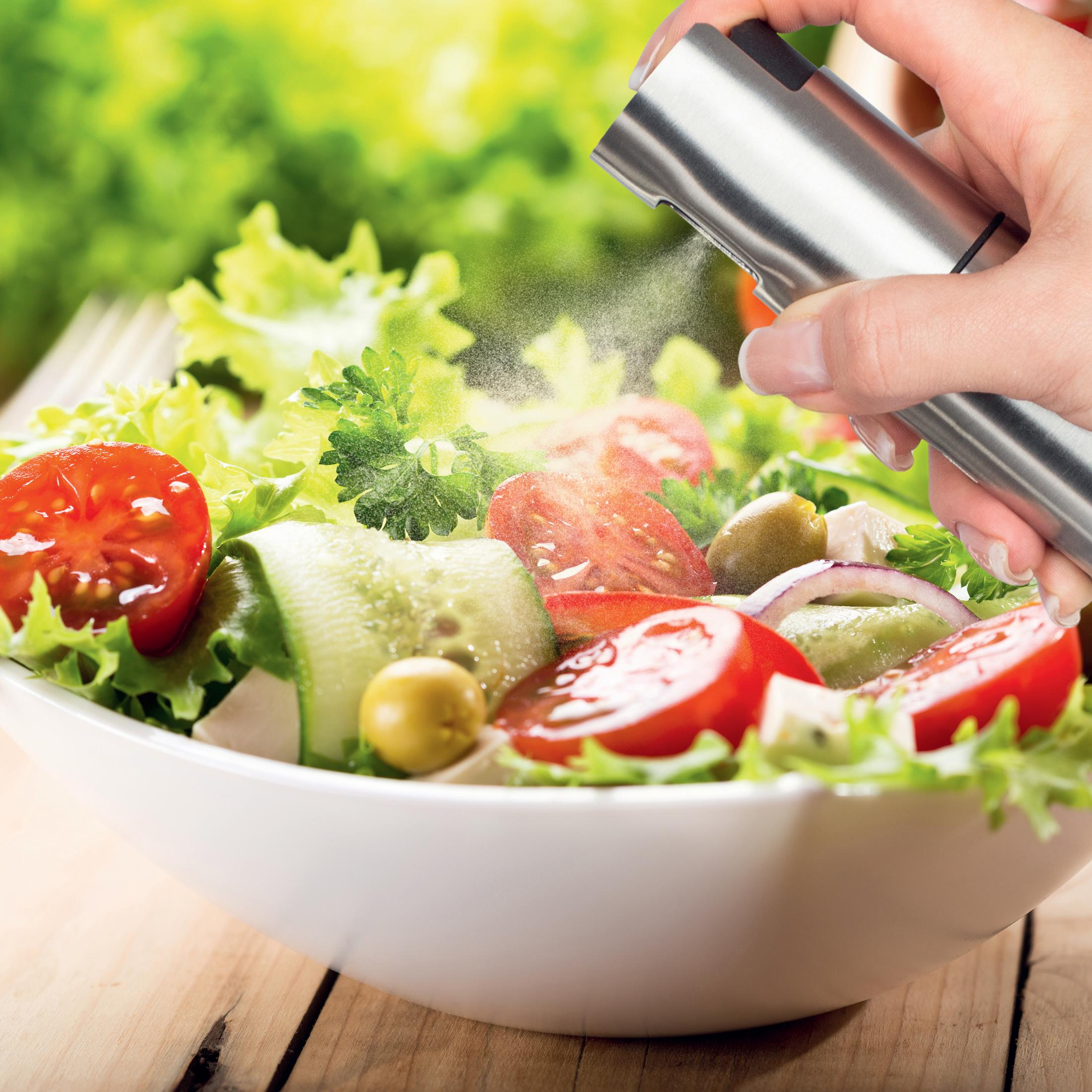 Eine Hand sprüht Öl auf einen frischen Salat mit Tomaten, Gurken und Oliven.