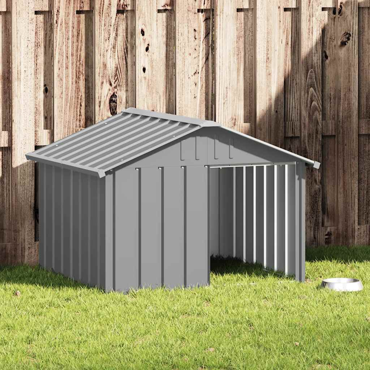 Graues Hundehäuschen aus Metall mit offenem Eingang vor einem Holzzaun und Gras.