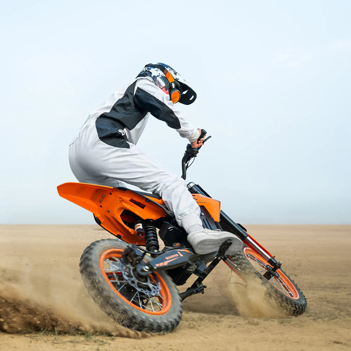Osoba w białym stroju i kasku jedzie pomarańczowym motocyklem crossowym, wzbijając kurz.