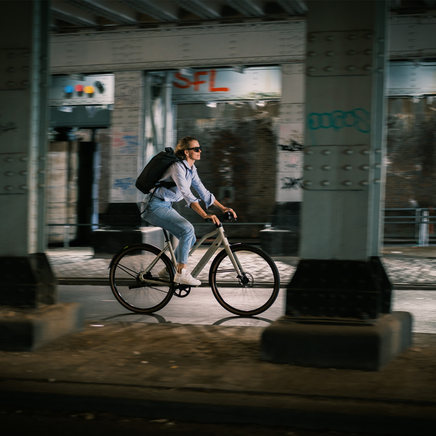 Eine Frau fährt nachts mit einem Fahrrad unter einer Brücke, beleuchtet von Laternen.