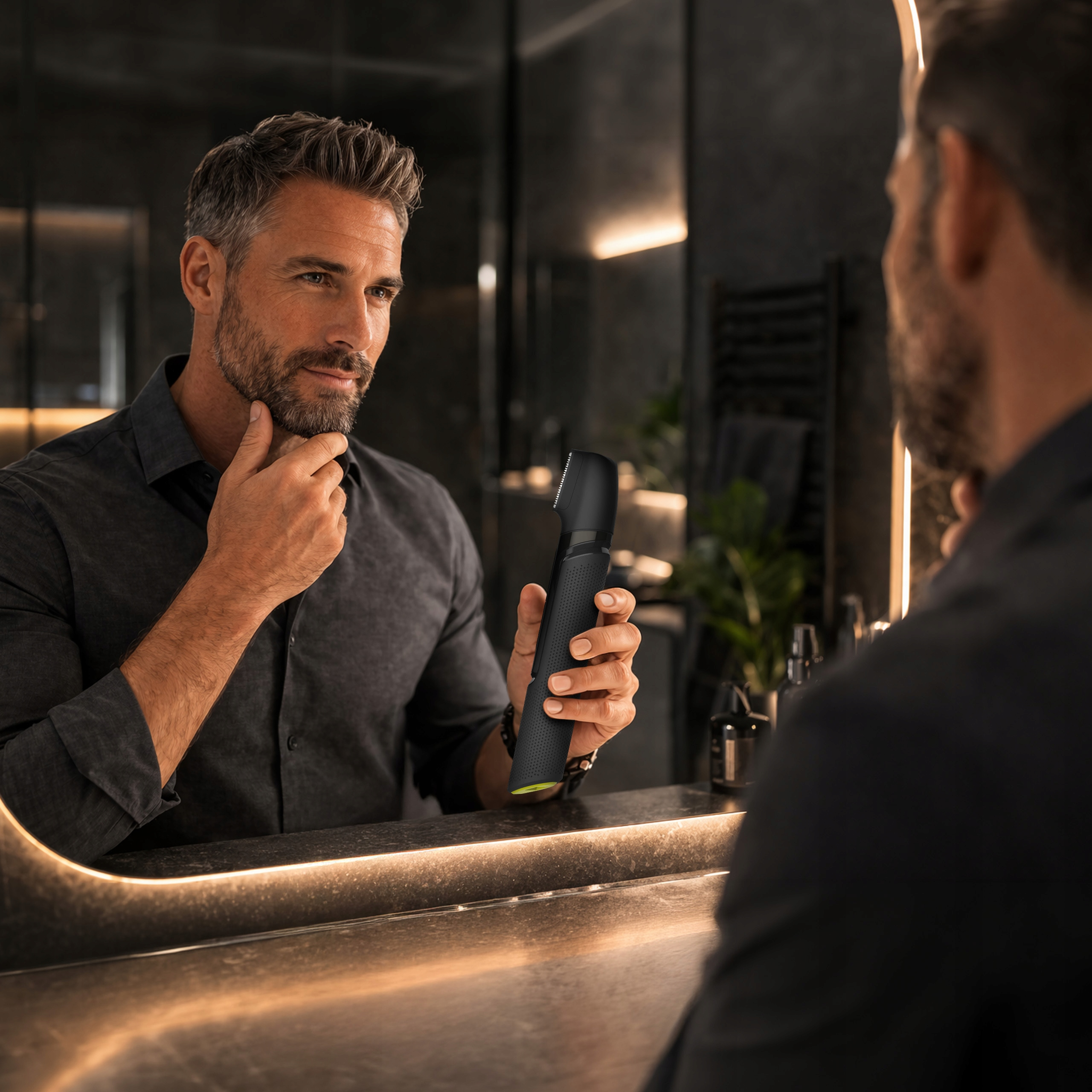 Mężczyzna z siwiejącymi włosami i brodą patrzy w lustro, trzyma czarne narzędzie.