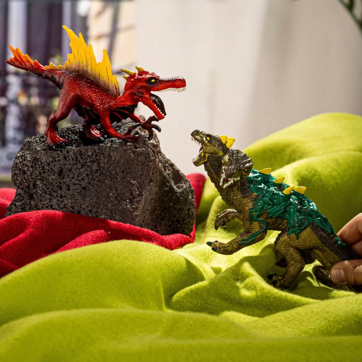 Rote und grüne Dinosaurier-Spielzeuge stehen sich auf einem Bett aus grünem und rotem Stoff gegenüber.
