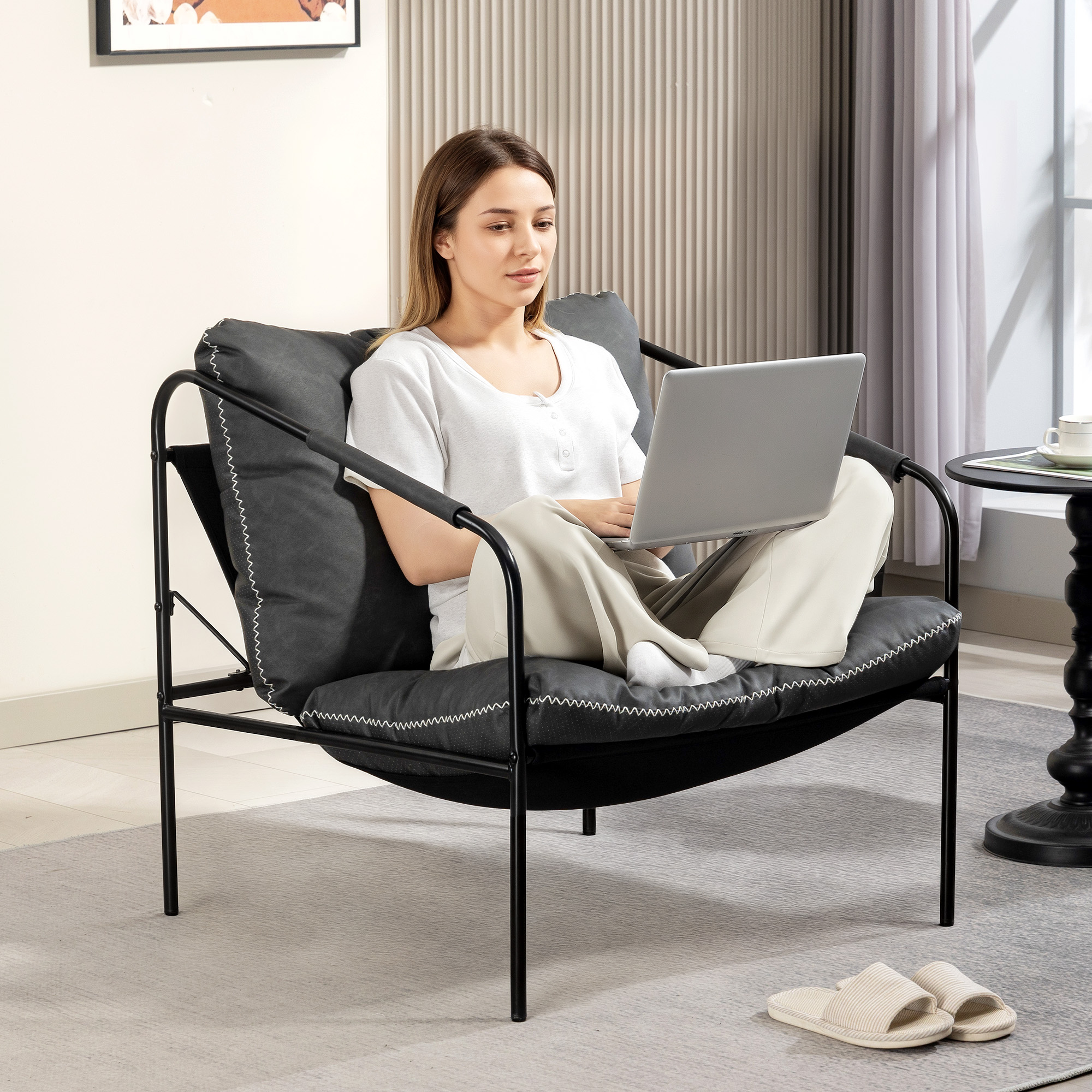 Una mujer se sienta en una silla gris oscuro usando una laptop. Una pequeña mesa negra con una taza está cerca.