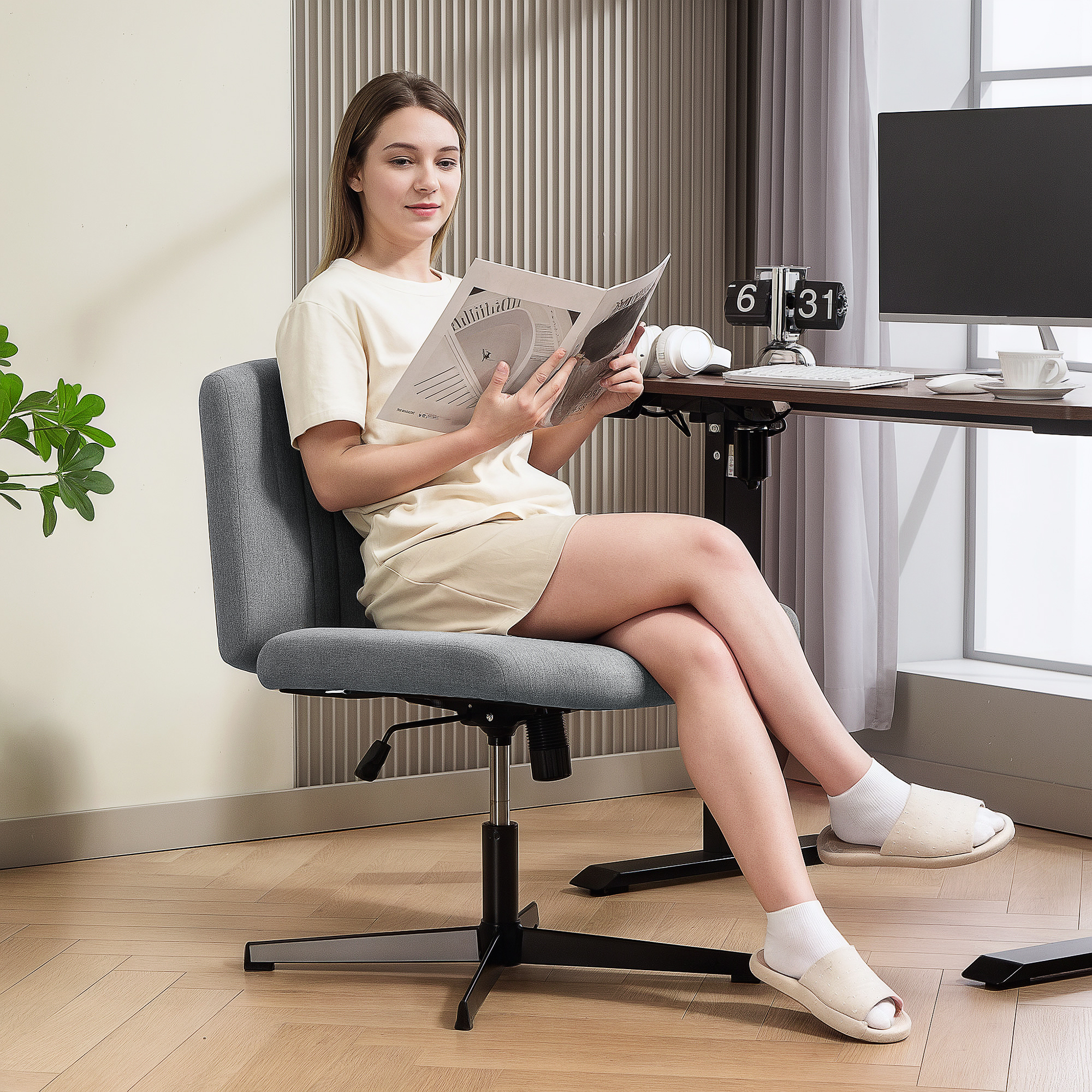 Una mujer sentada en silla de oficina lee una revista. Un escritorio con computadora al fondo.