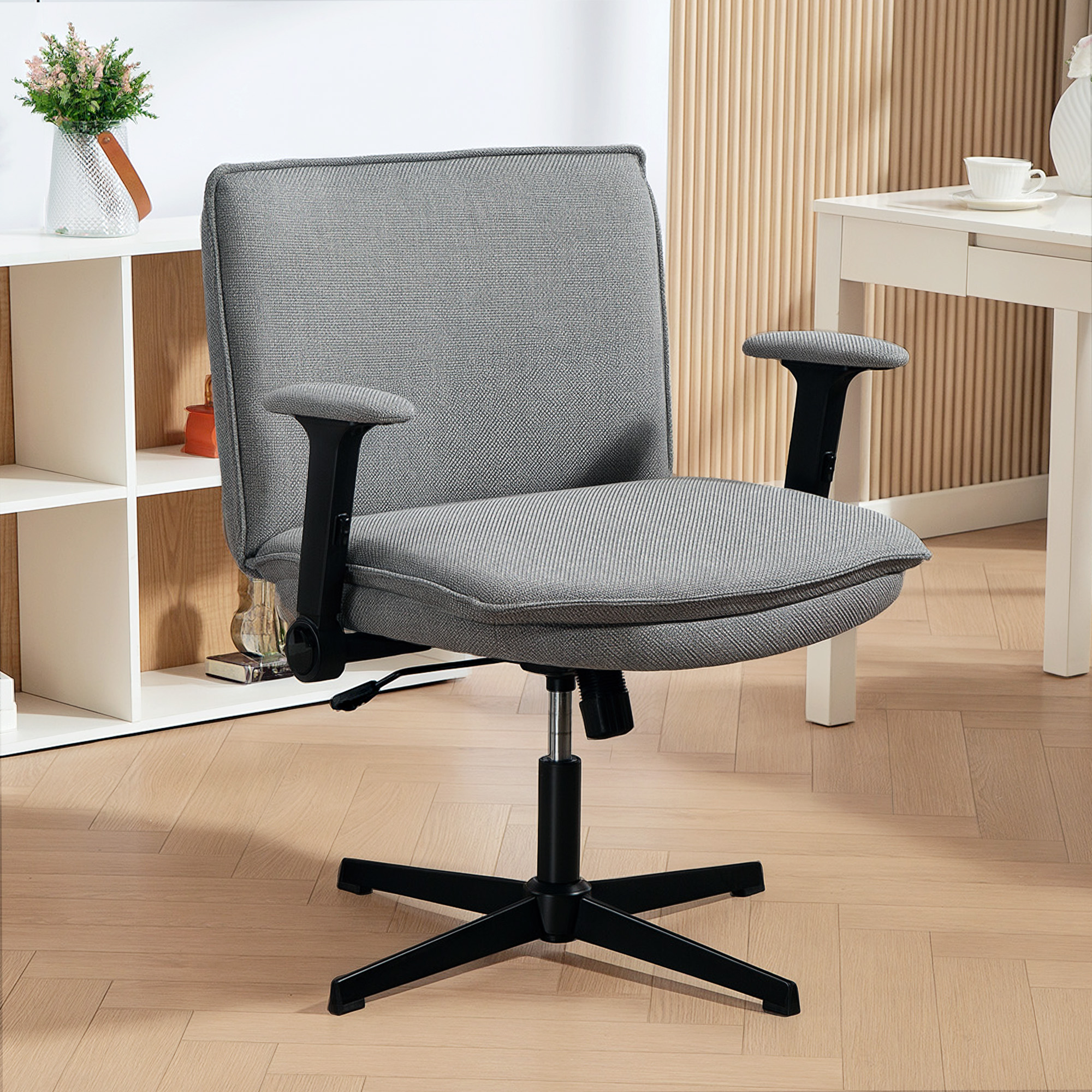 Una silla de oficina gris con reposabrazos y base negros se encuentra sobre un suelo de madera clara. Un estante blanco y una mesa están al fondo.