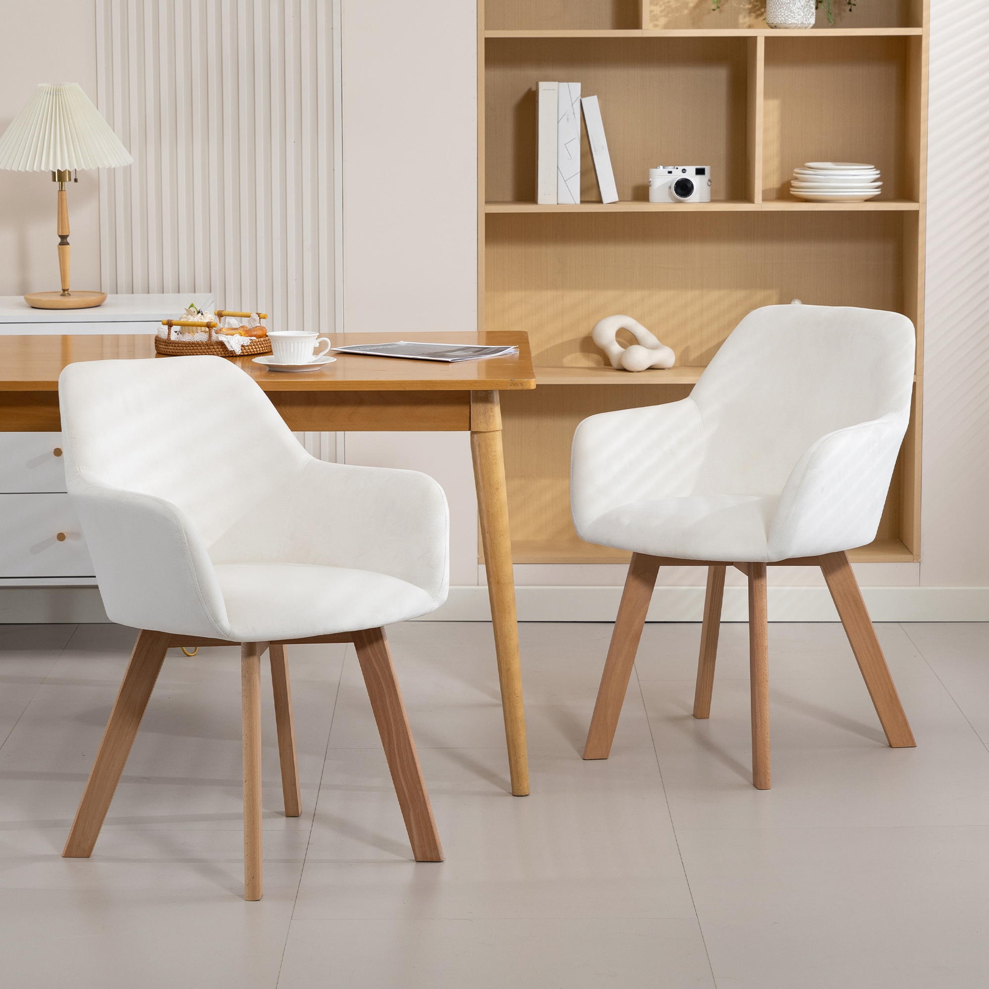 Dos sillas blancas con patas de madera junto a una mesa de madera. Una estantería tiene libros.