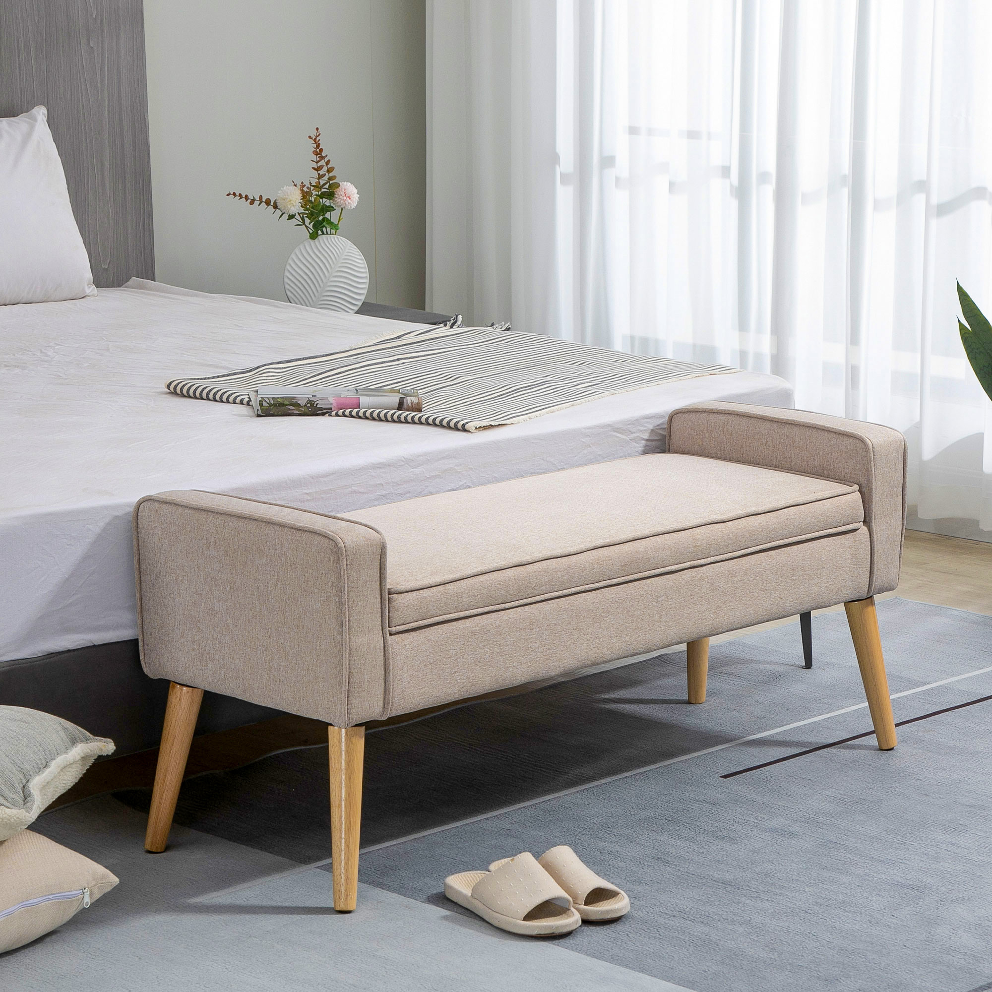 Un banco tapizado beige con patas de madera se sienta frente a una cama con ropa blanca y manta a rayas.