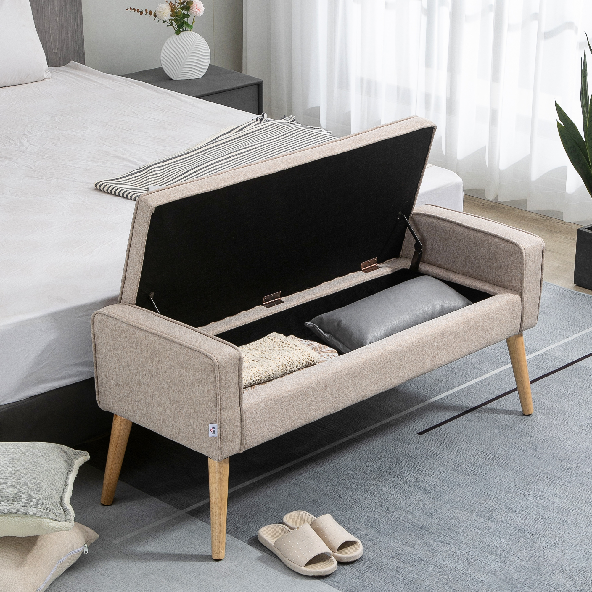 Banco de almacenamiento beige con tapa abierta junto a una cama. Cojines y mantas dentro.