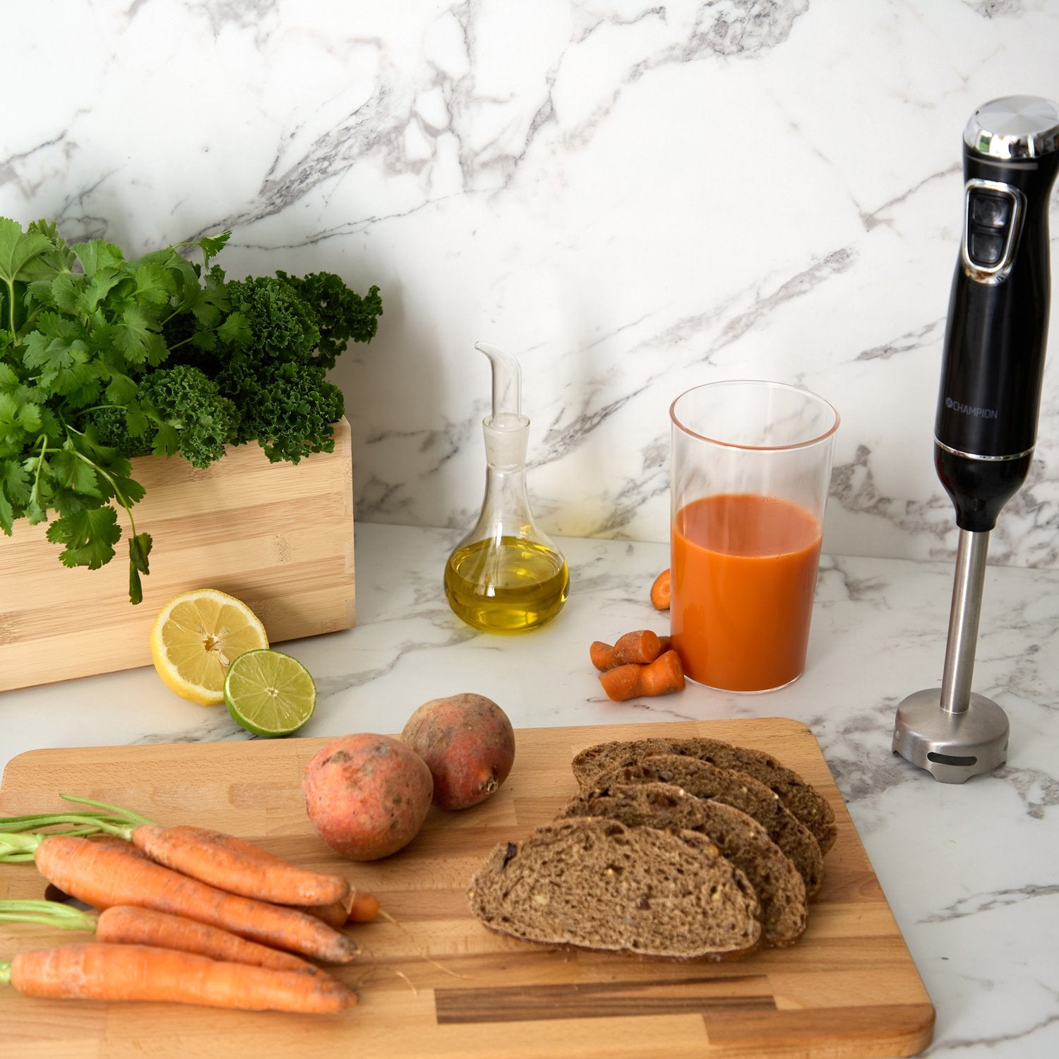 A hand blender stands beside a glass of carrot juice. Ingredients like carrots, bread, and herbs are ready.