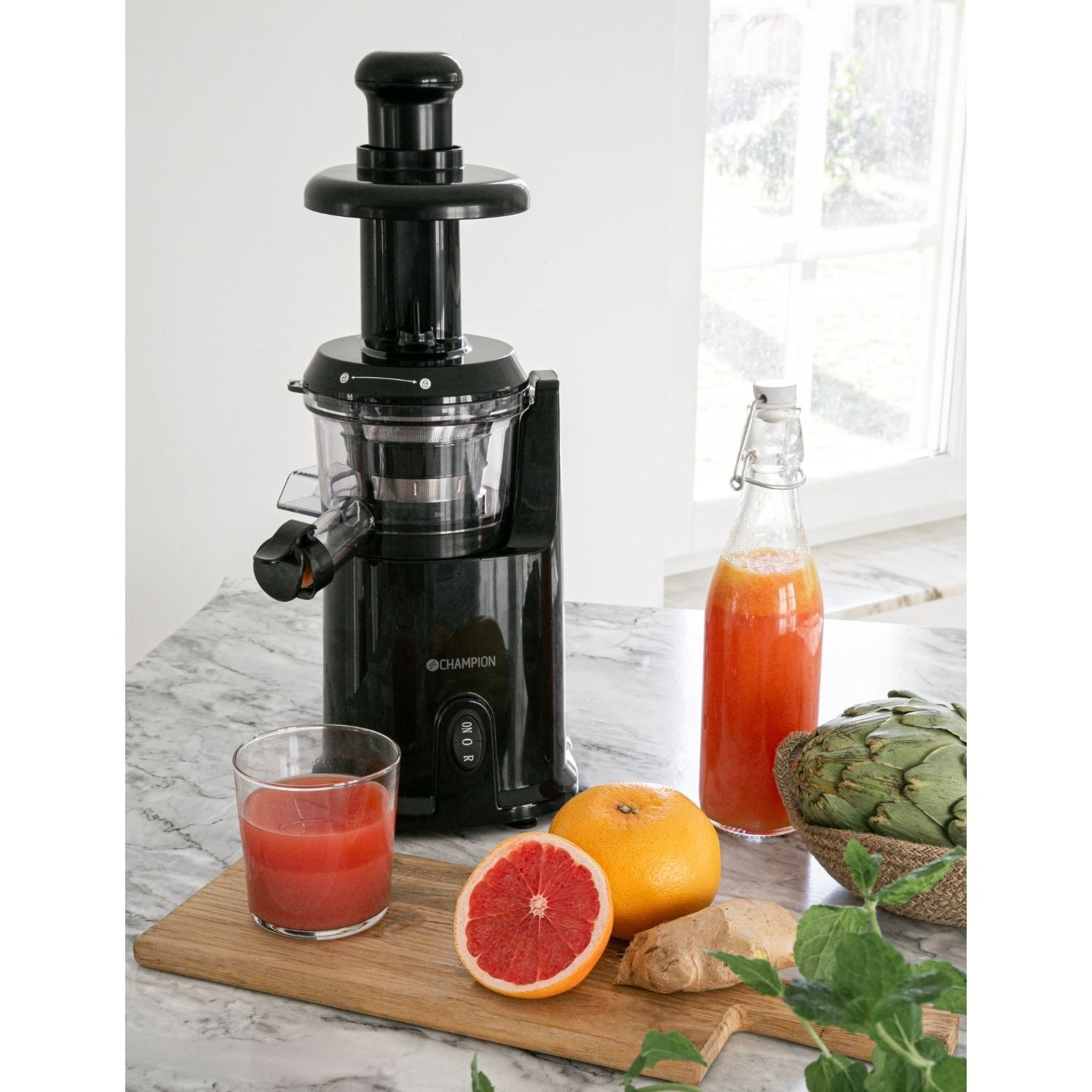 Black juicer with grapefruit, juice bottle, artichoke, ginger, and mint on a marble counter.
