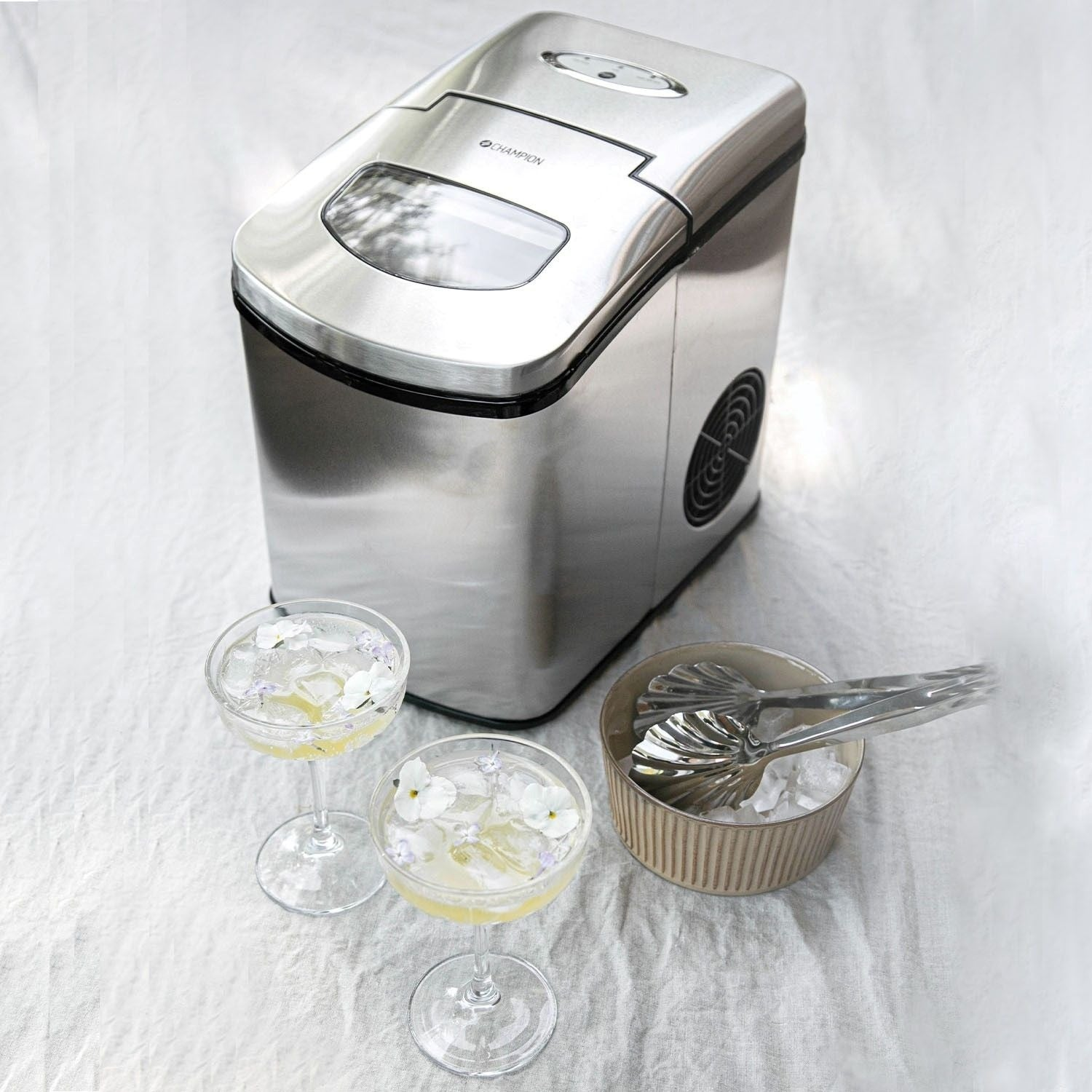 A silver ice maker stands next to two cocktails adorned with flowers.