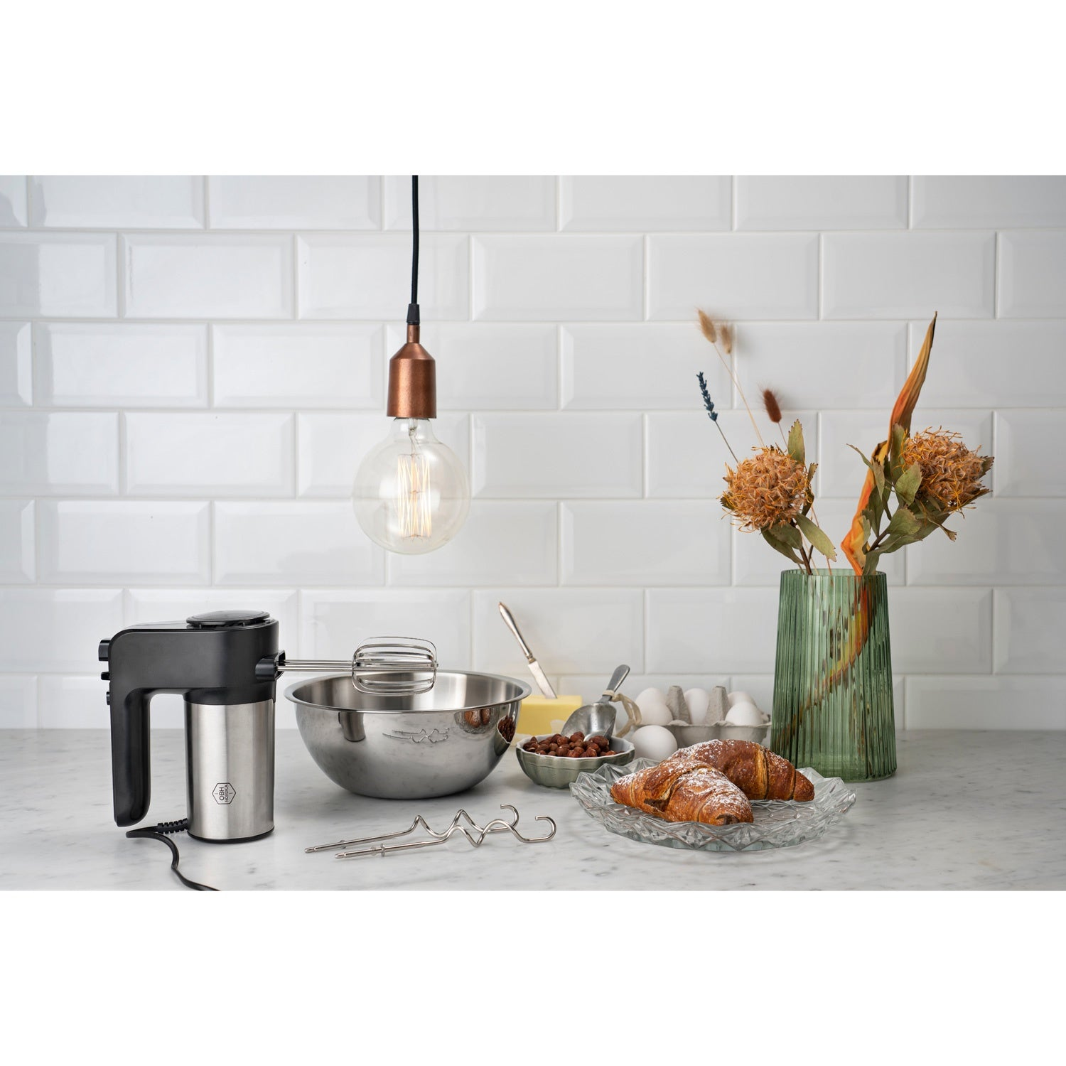 Hand mixer with bowl, croissants, eggs, and flowers in vase on countertop.