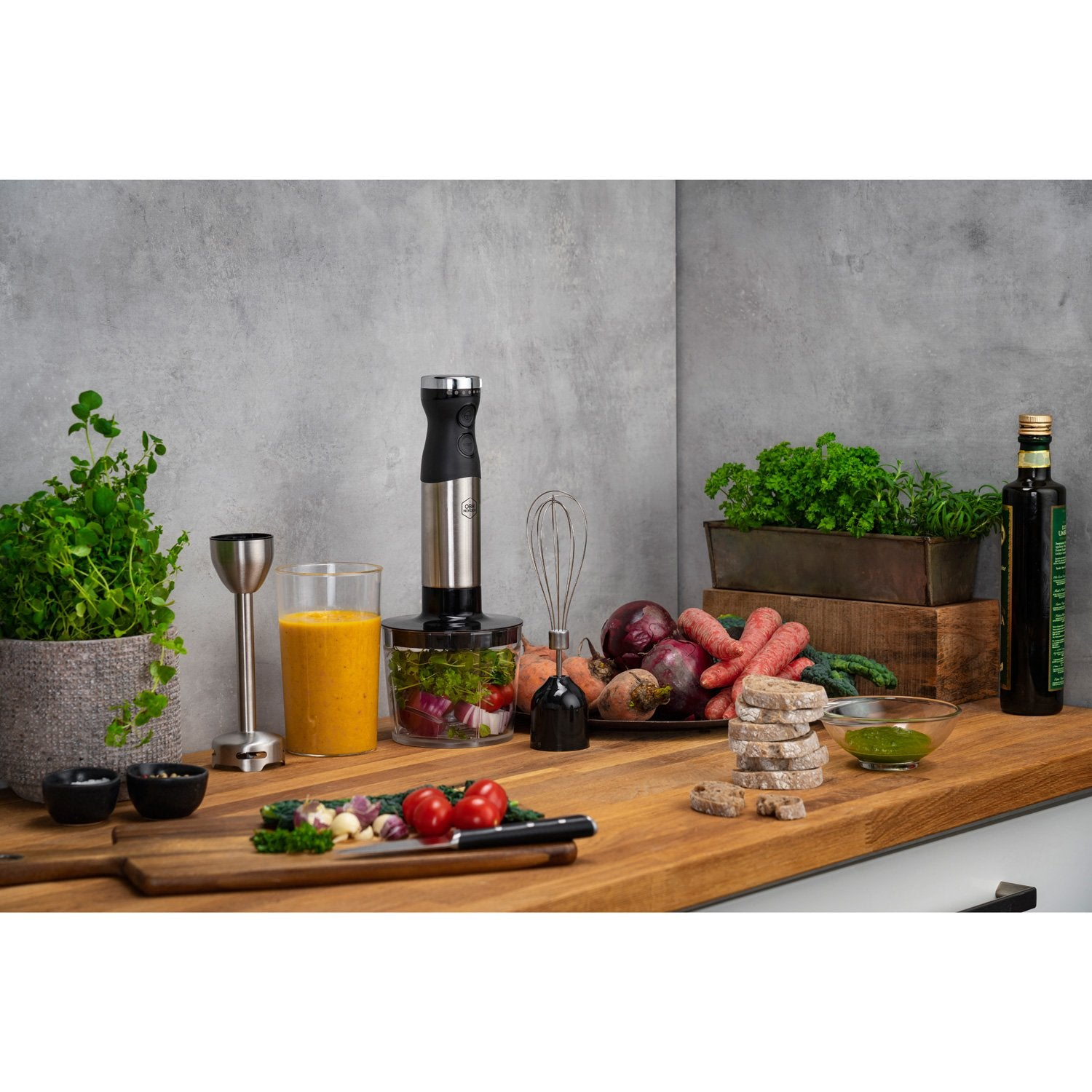 An immersion blender beside a glass of juice and fresh vegetables on a wooden counter.