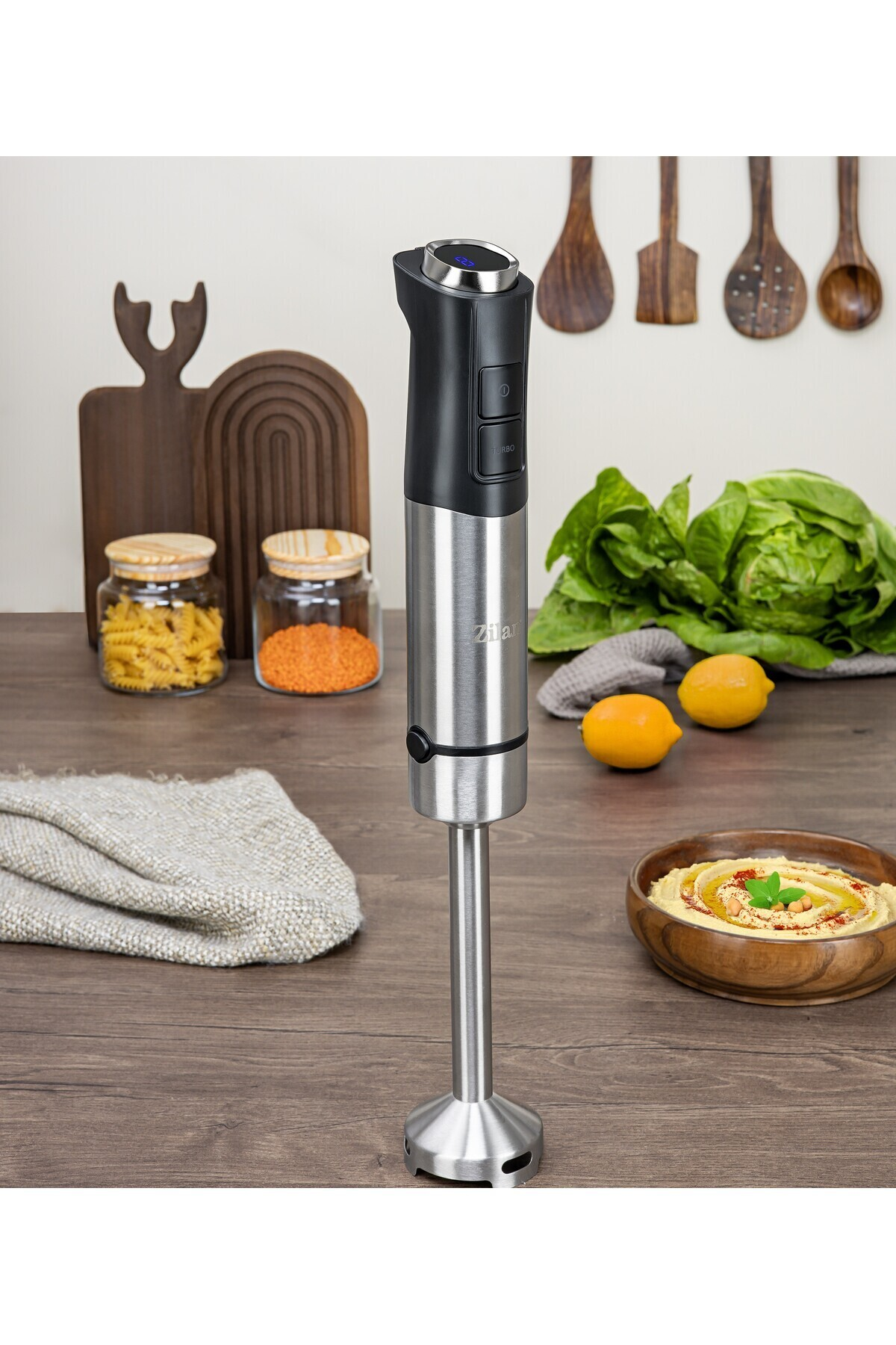 Ahşap bir masanın üzerinde duran el blenderı, yanında humus ve marul.