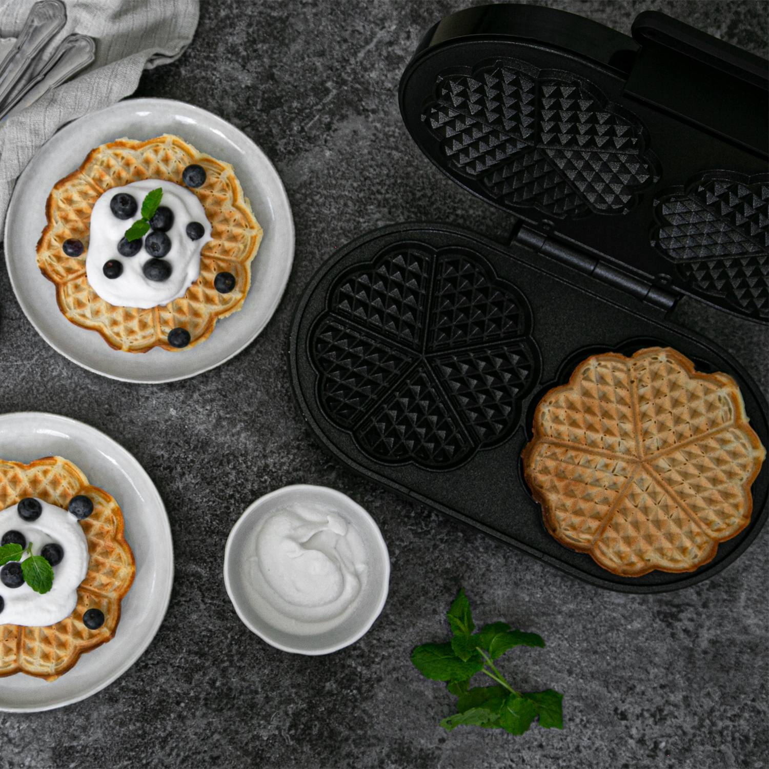 Waffle maker with waffles, cream, blueberries, and mint on a dark surface.