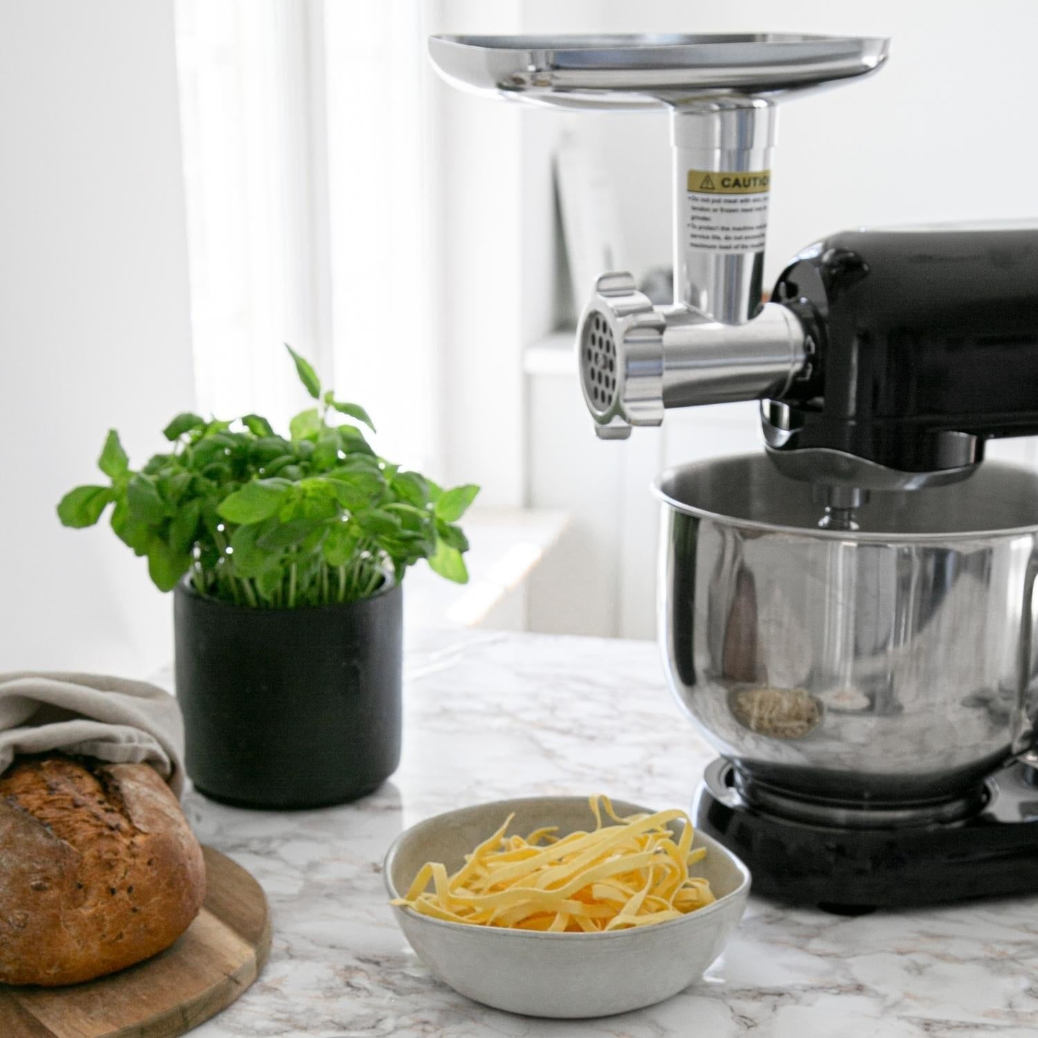 Stand mixer with meat grinder attachment, bread, herb pot, and pasta bowl.