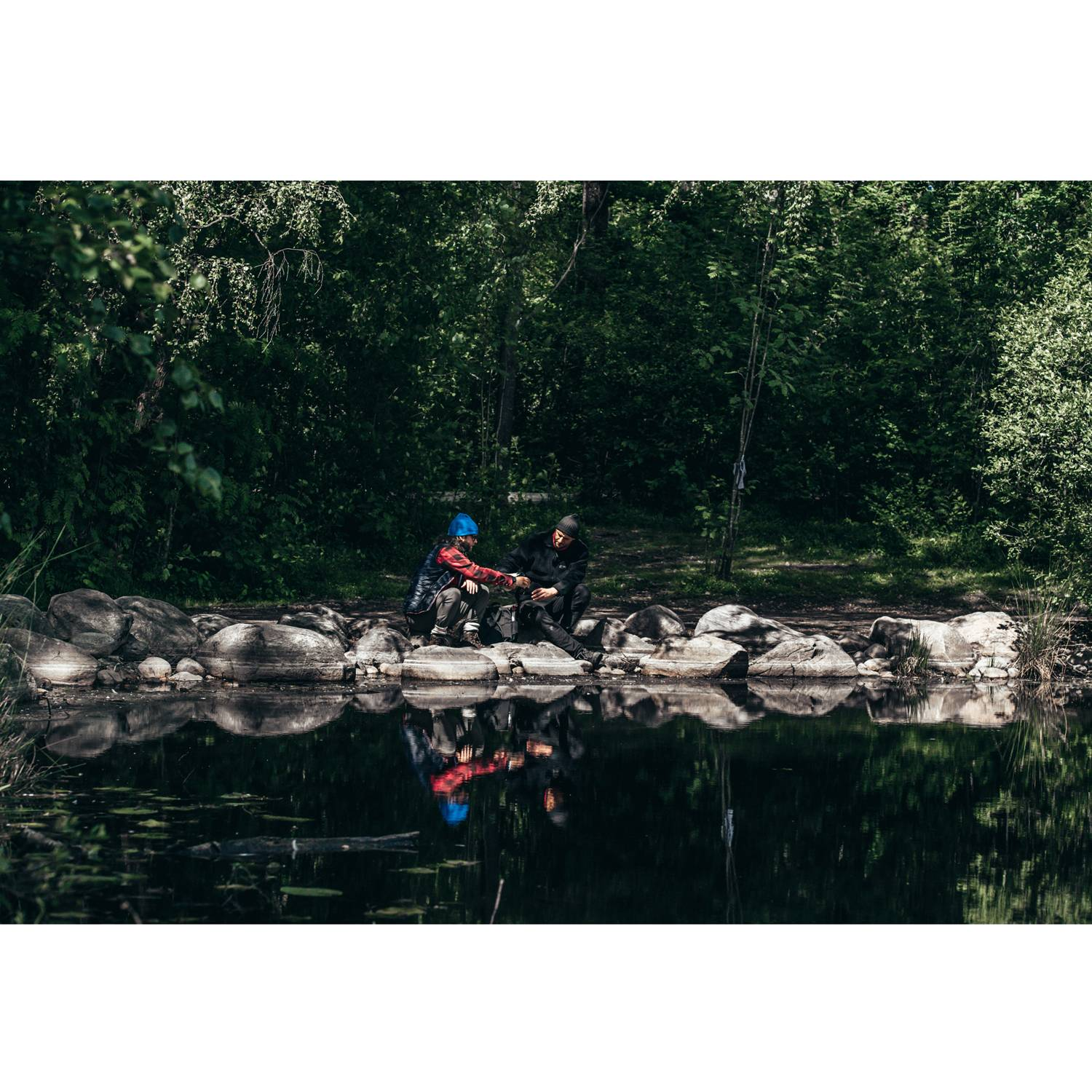 Twee mensen zitten op rotsen aan een donker meer, omringd door weelderige groene bomen.