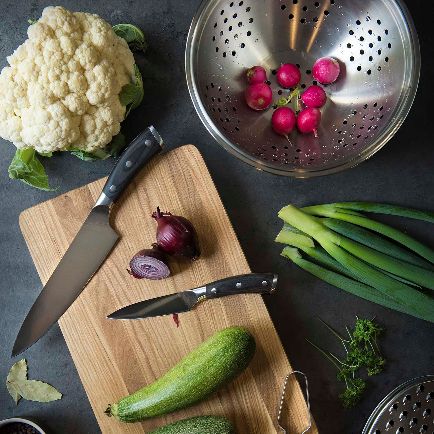 Bloemkool, uien, radijsjes, lente-uitjes, courgette en kruiden op een donkere tafel.