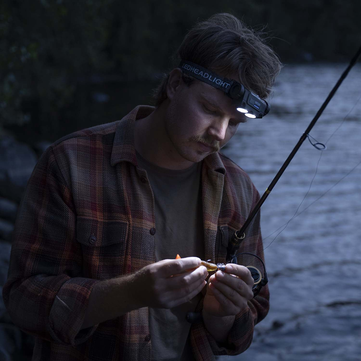Man with headlight attaches lure to fishing rod by river at dusk.
