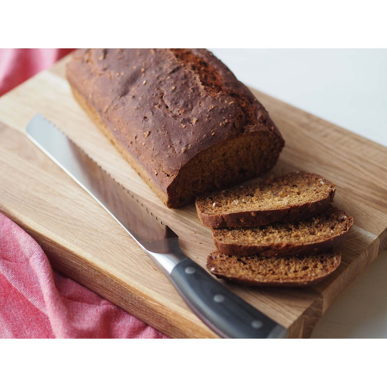 Een donker brood met drie sneetjes op een houten plank naast een mes.