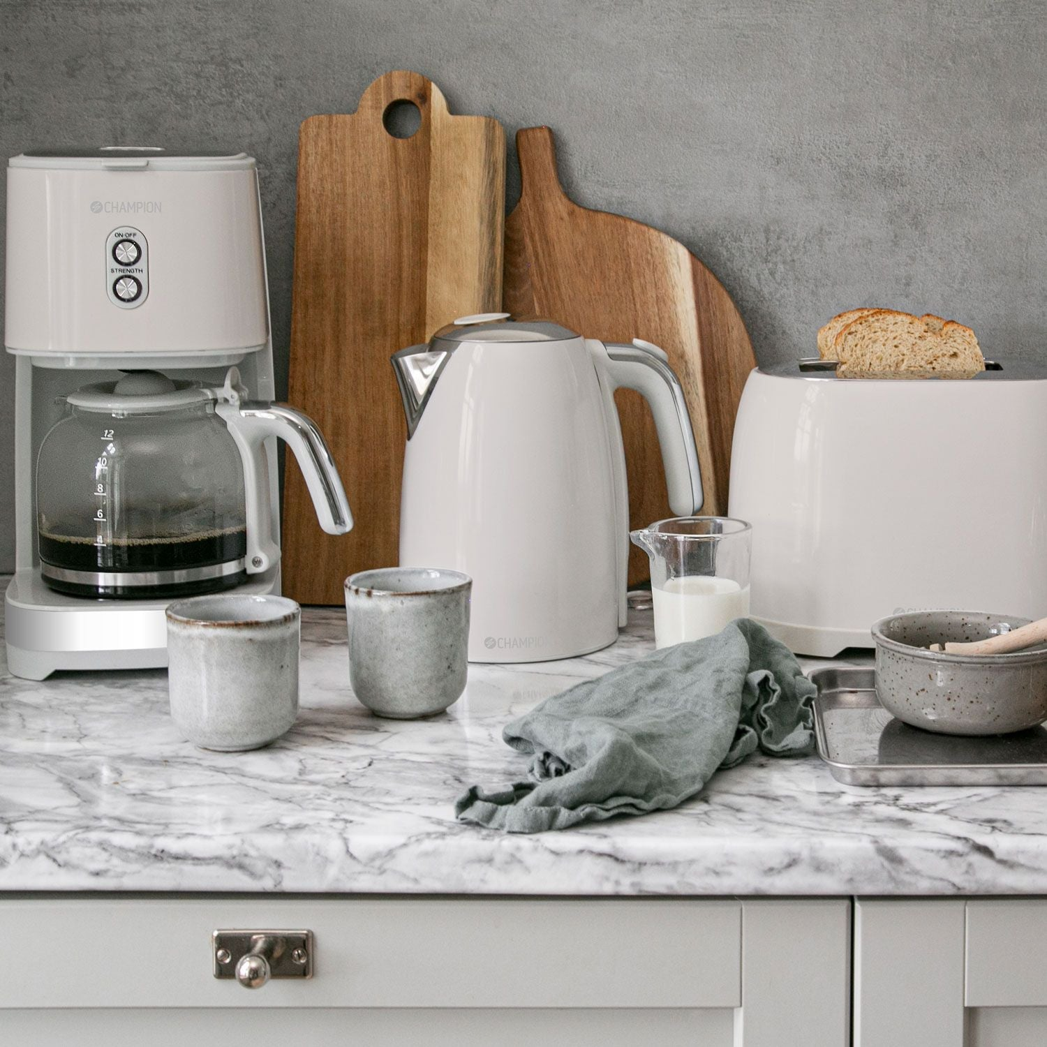 Wit koffiezetapparaat, waterkoker, broodrooster met brood, kopjes en houten snijplanken.