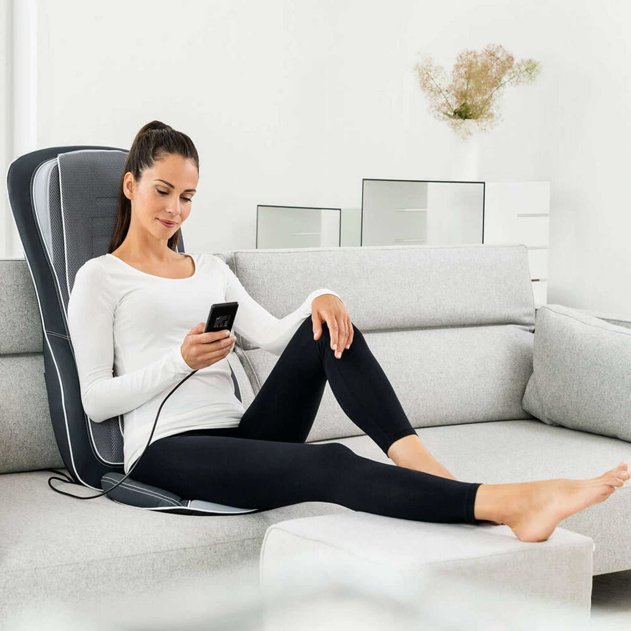 Eine Frau sitzt auf einem Sofa und benutzt ein Massagegerät, während sie ihr Handy hält.