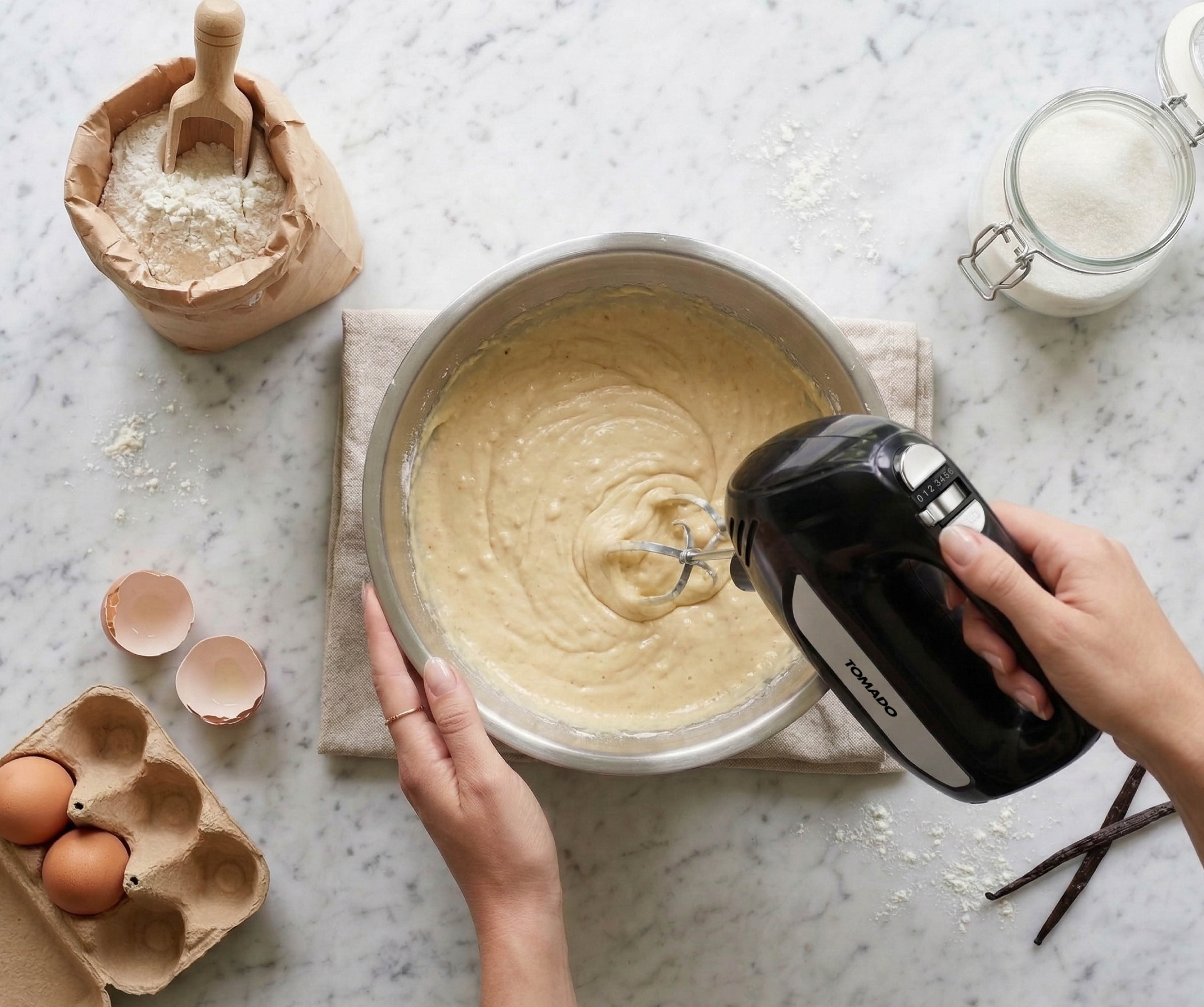 A hand uses a black hand mixer to stir batter in a metal bowl. Ingredients nearby.