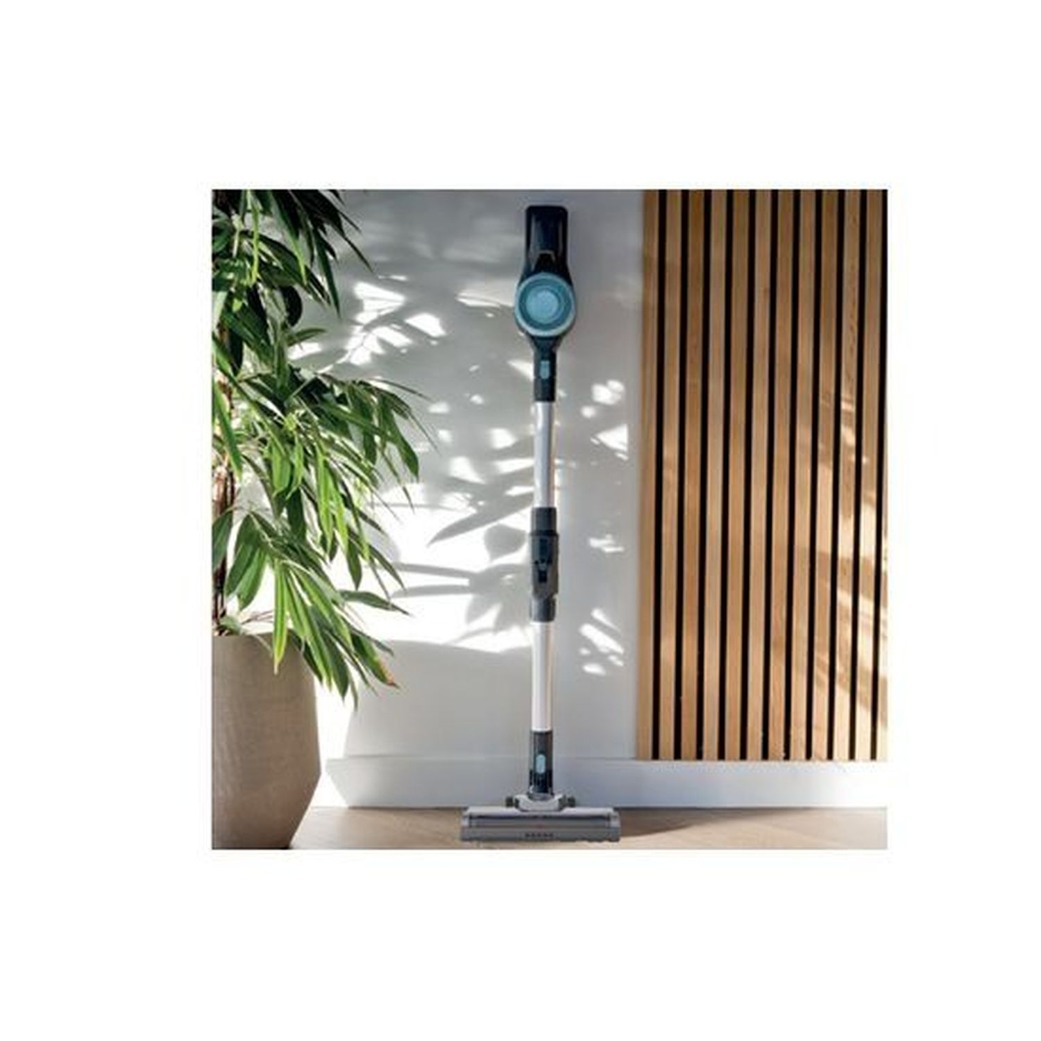 A sleek, upright vacuum cleaner stands before vertical wood paneling and a potted plant.