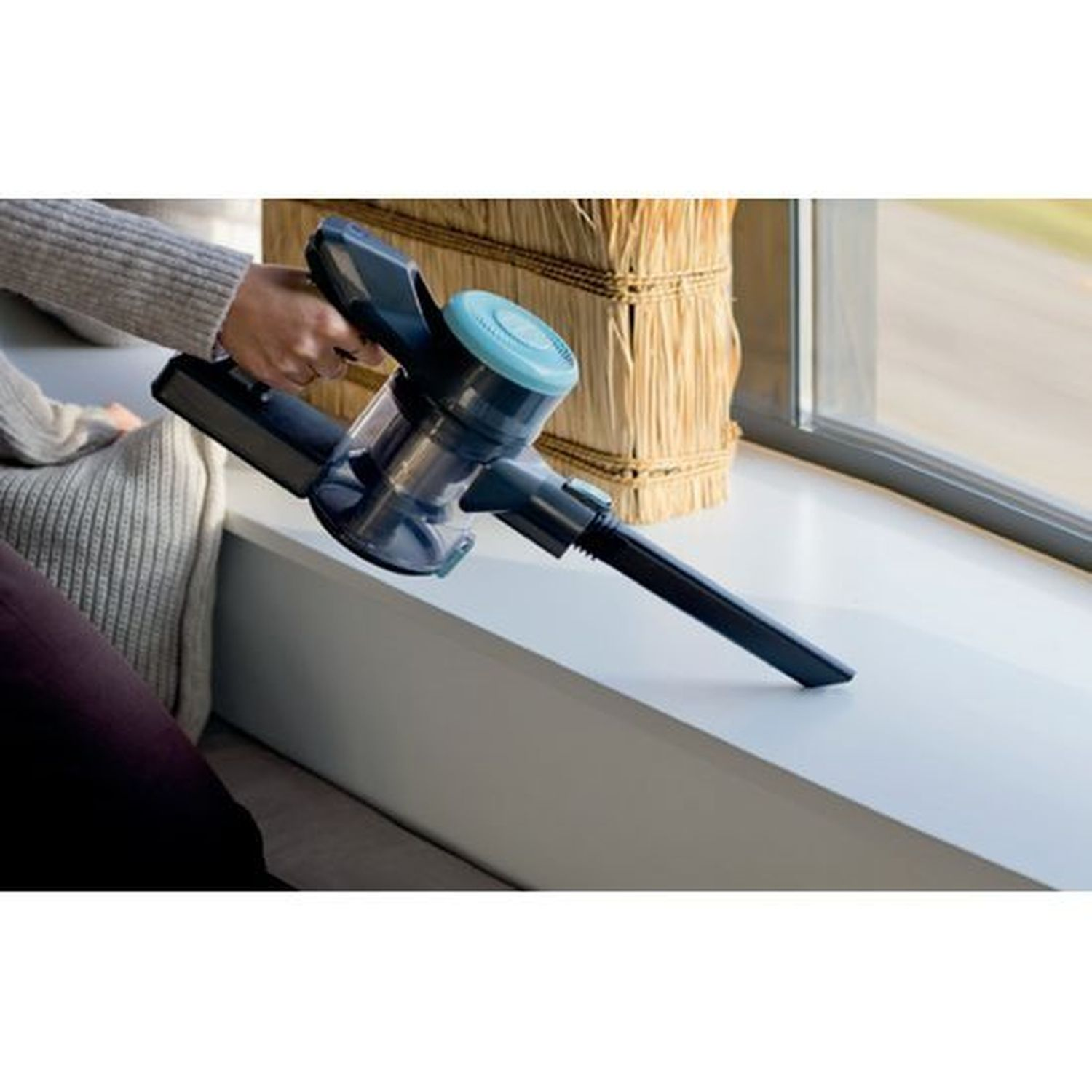 A hand holds a blue and black handheld vacuum cleaner on a windowsill.
