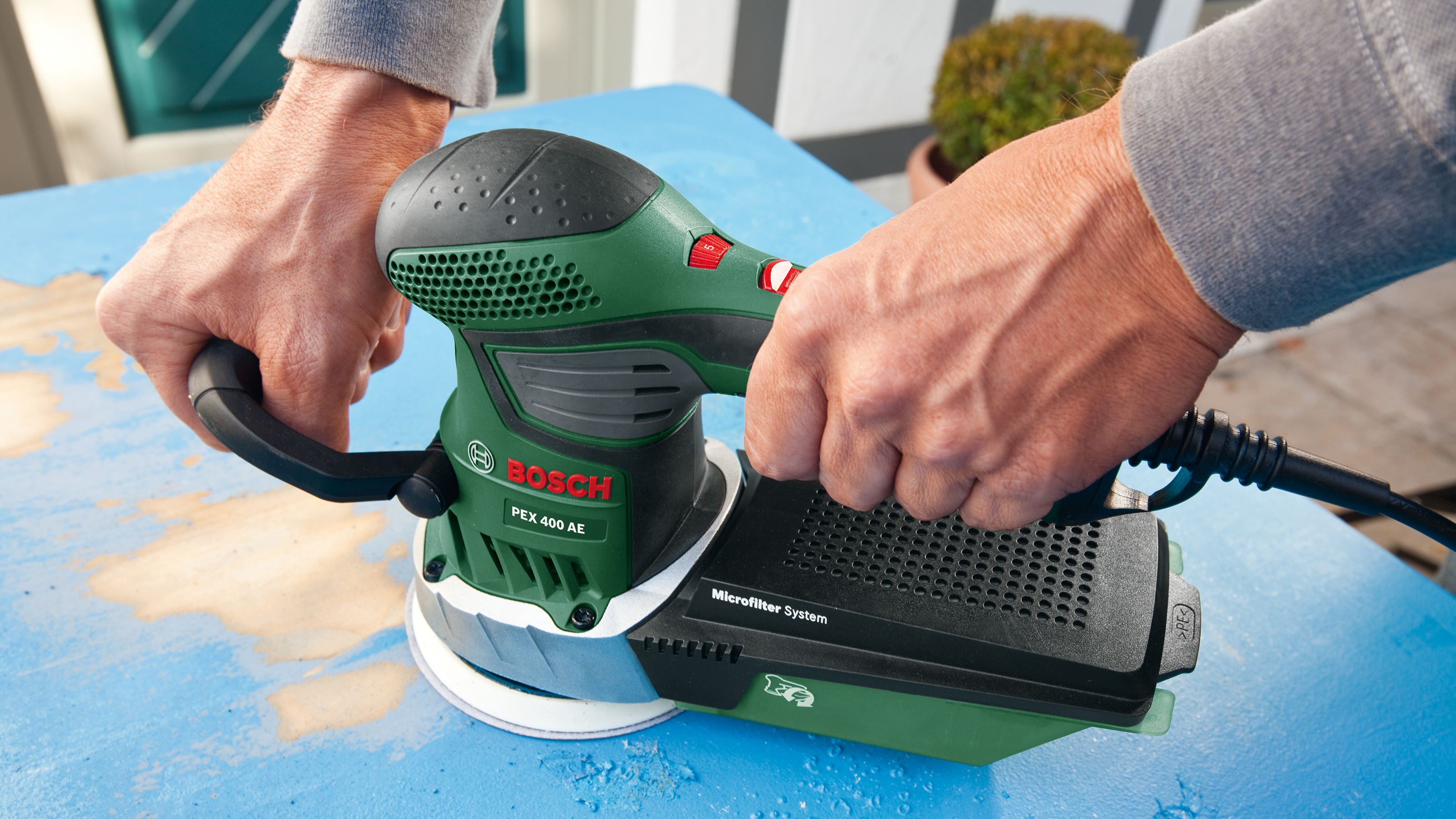 Une ponceuse Bosch verte est tenue par des mains sur une surface bleue.