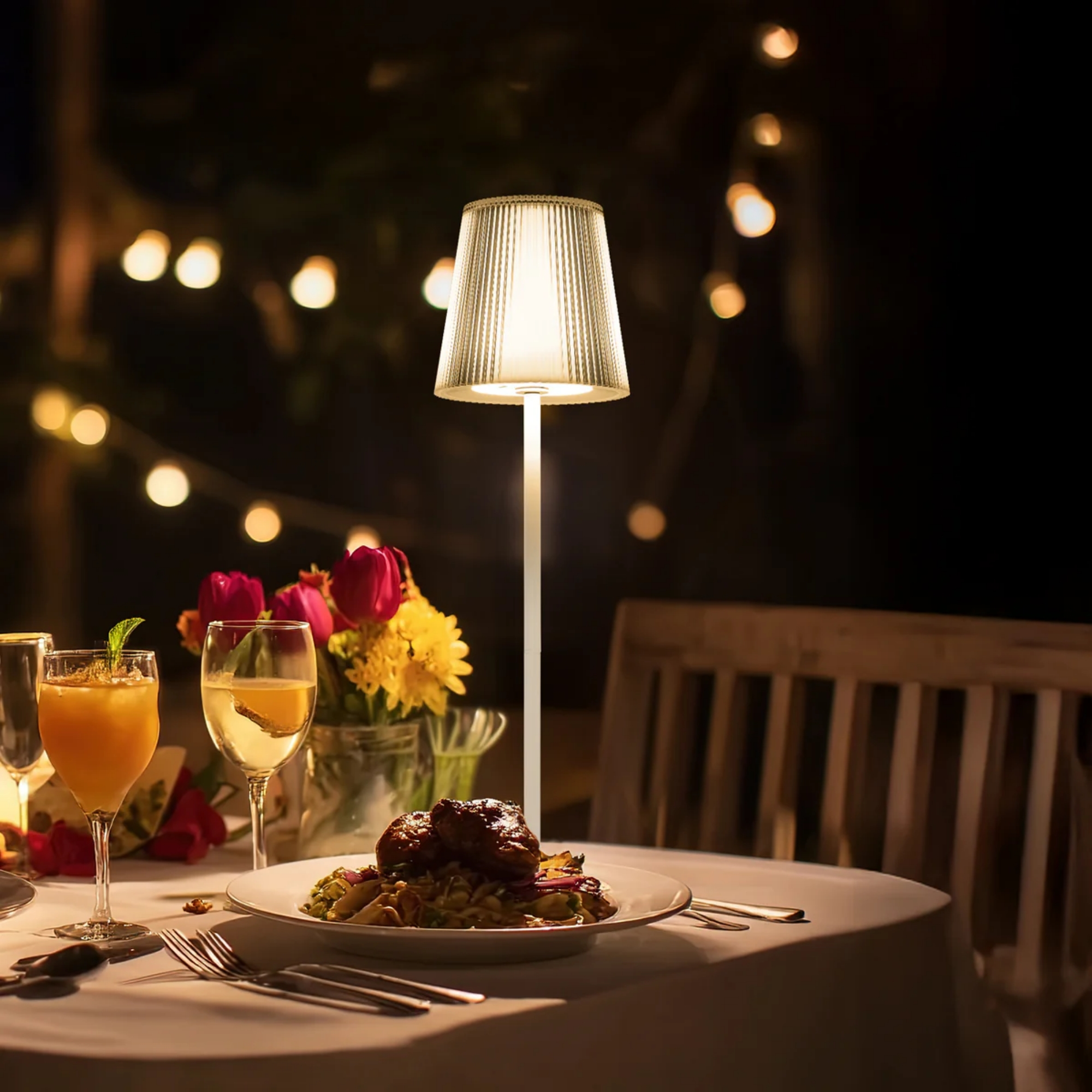 Ein beleuchteter Tisch mit Essen, Getränken und Blumen im Freien bei Nacht.