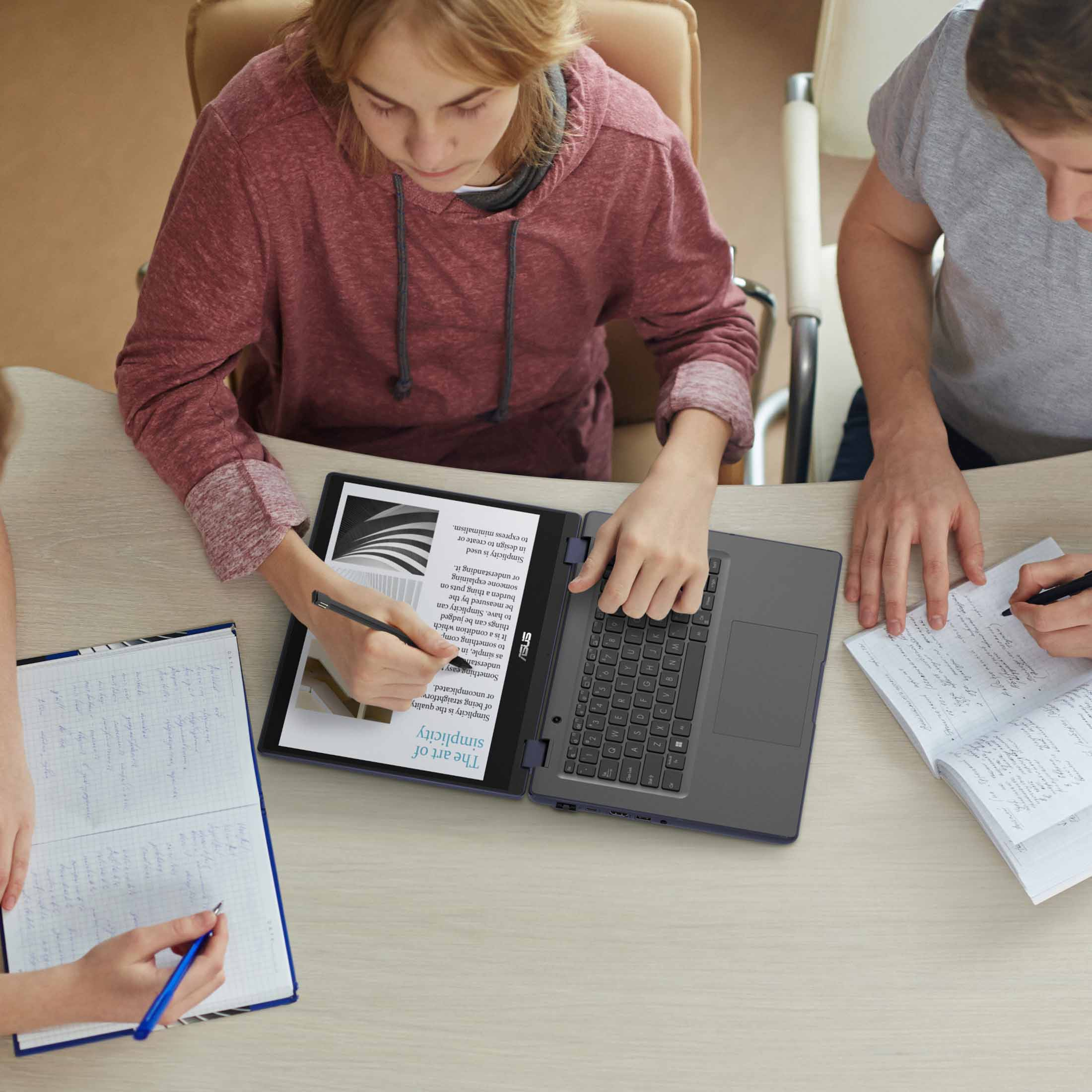 Drei Personen lernen an einem Holztisch, eine benutzt einen Laptop, zwei schreiben in Notizbücher.
