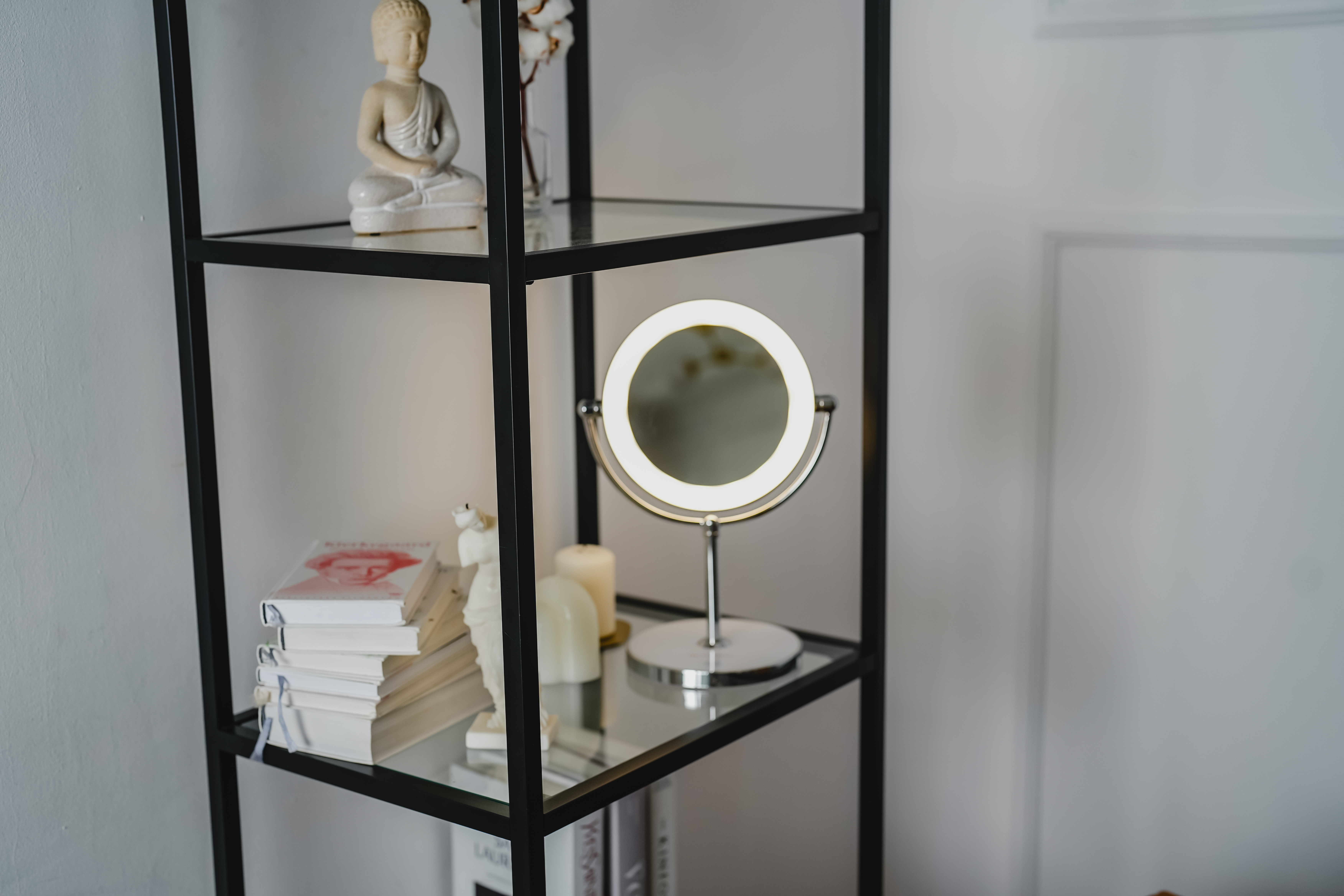 Ein schwarzes Metallregal zeigt Dekoartikel: Buddha-Statue, Bücher, Kerzen und ein beleuchteter Spiegel.