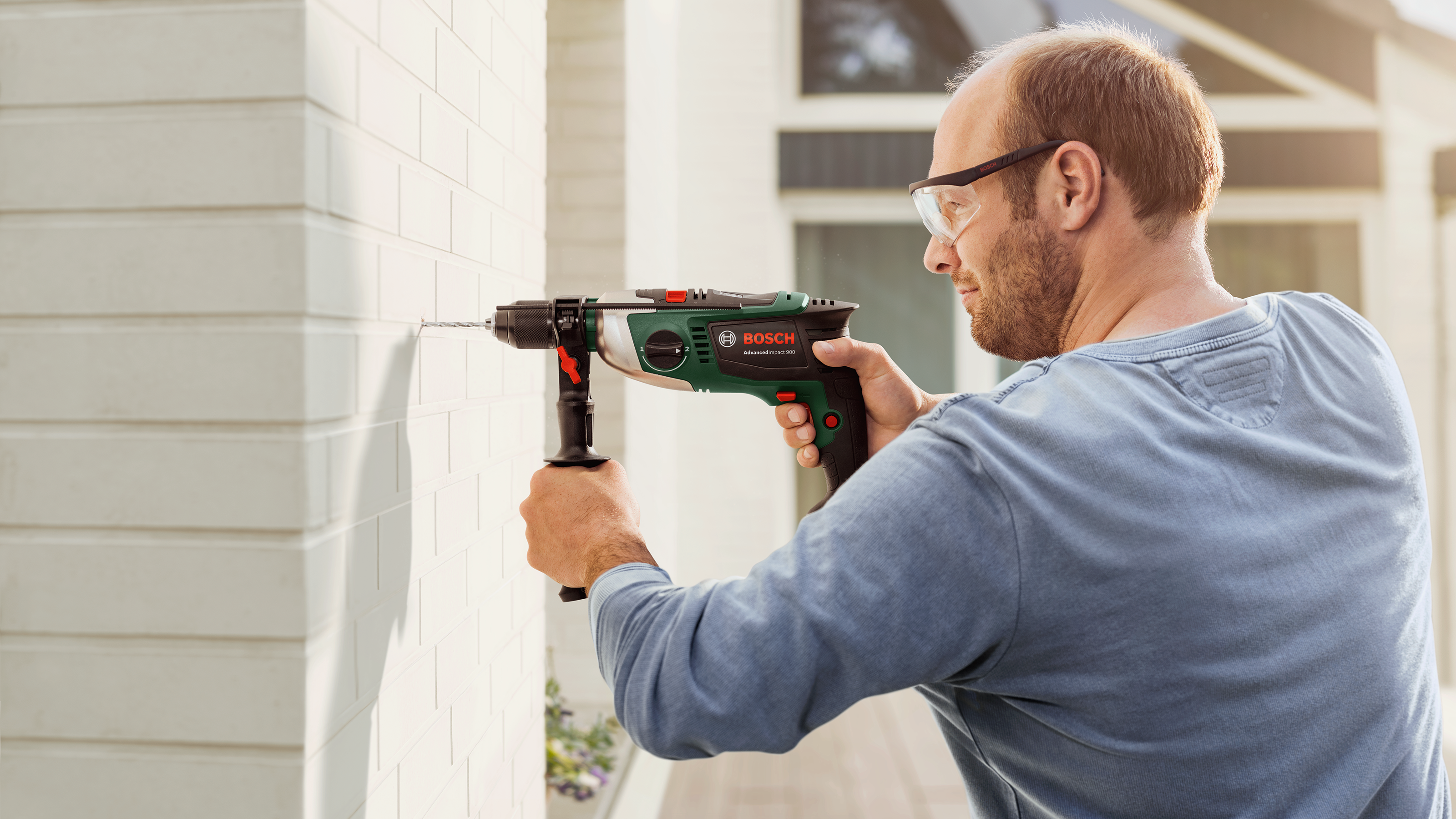 Mężczyzna wierci dziurę w białej ścianie; nosi okulary i niebieską koszulę. Narzędzie zielono-czarne.
